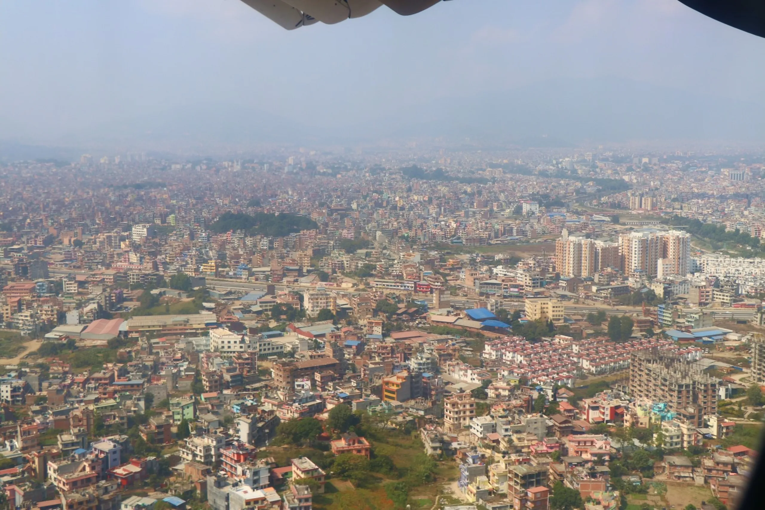 A stunning aerial window-seat photograph taken from inside a Buddha Air passenger aircraft on approach or departure over Kathmandu Valley, Nepal - looking down through the slightly curved aircraft window frame at the vast densely packed urban sprawl of Kathmandu city spread across the valley floor, thousands of multi-story brick and concrete residential and commercial buildings in shades of terracotta, white, grey and blue rooftops extending to the horizon in every direction, a low hill with dense tree cover rising in the middle left of the cityscape, several larger modern buildings and construction sites visible to the right, the entire city blanketed in a thin layer of urban smog and haze under a grey-white overcast sky, aerial view Kathmandu Valley Nepal, Kathmandu city from plane Nepal, Buddha Air Nepal approach Kathmandu, Tribhuvan International Airport approach Nepal, Kathmandu aerial photography, Nepal capital city from above flight