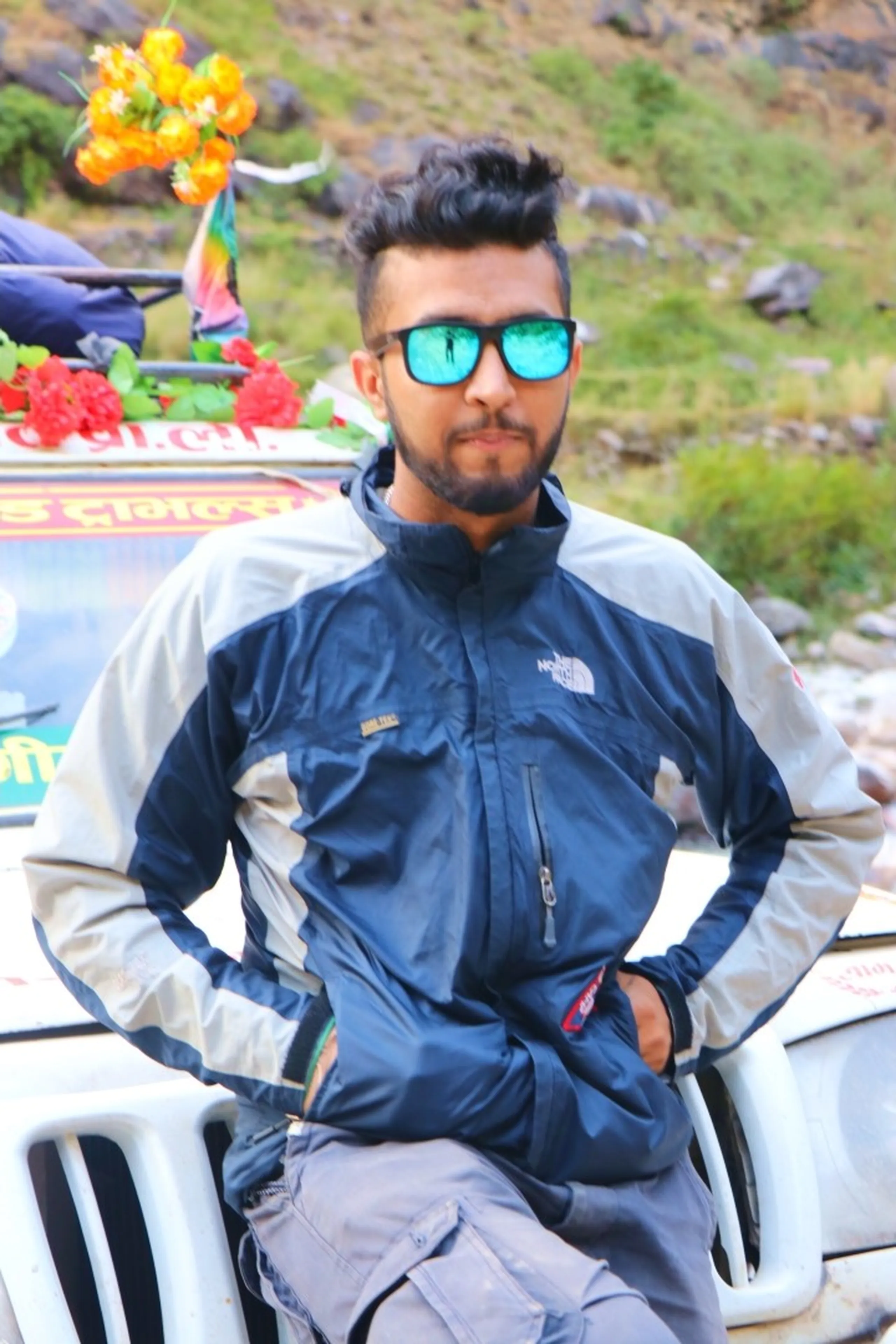 A stylish young South Asian male trekker posing with arms behind his back in front of a Nepali jeep decorated with red artificial flowers, wearing a two-tone navy and silver-grey North Face windbreaker jacket and grey cargo pants, sporting teal mirrored rectangular sunglasses and a neatly quiffed black hairstyle with a short beard, rocky green mountain hillside visible in the background, Nepali Devanagari signage on the vehicle, cool trekking fashion Nepal, North Face gear Himalayan trek, Shey Phoksundo trek style, adventure travel Nepal young traveler