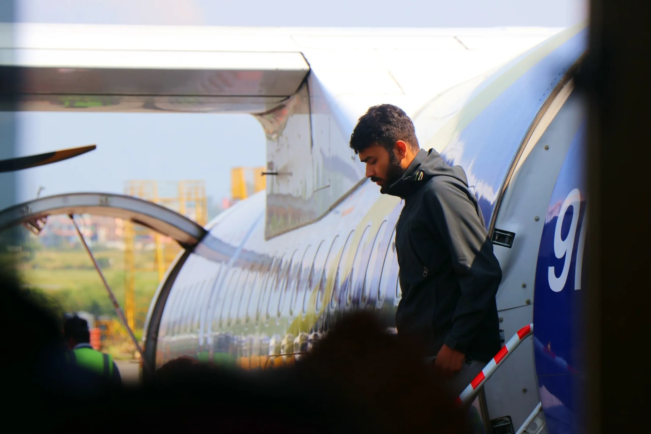 A candid and atmospheric telephoto portrait of a bearded young South Asian male trekker stepping down the aircraft stairs of a Buddha Air plane at Kathmandu's Tribhuvan International Airport, captured in profile through the aircraft window or terminal glass - wearing a dark charcoal zip-up hooded softshell jacket with headphones hanging around his neck, head bowed slightly as he descends the air-stair steps, yellow airport ground equipment and construction cranes visible in the blurred background, the aircraft's blue and white fuselage with a partial registration number "9I" or "9N" just visible at the right edge, a genuinely candid and reflective end-of-journey moment, candid trekker deplaning Kathmandu Nepal, Tribhuvan airport Nepal arrival candid, trekker returning Kathmandu Nepal Shey Phoksundo, Nepal domestic flight arrival photography, Buddha Air Nepal Kathmandu landing, post trek return Kathmandu Nepal