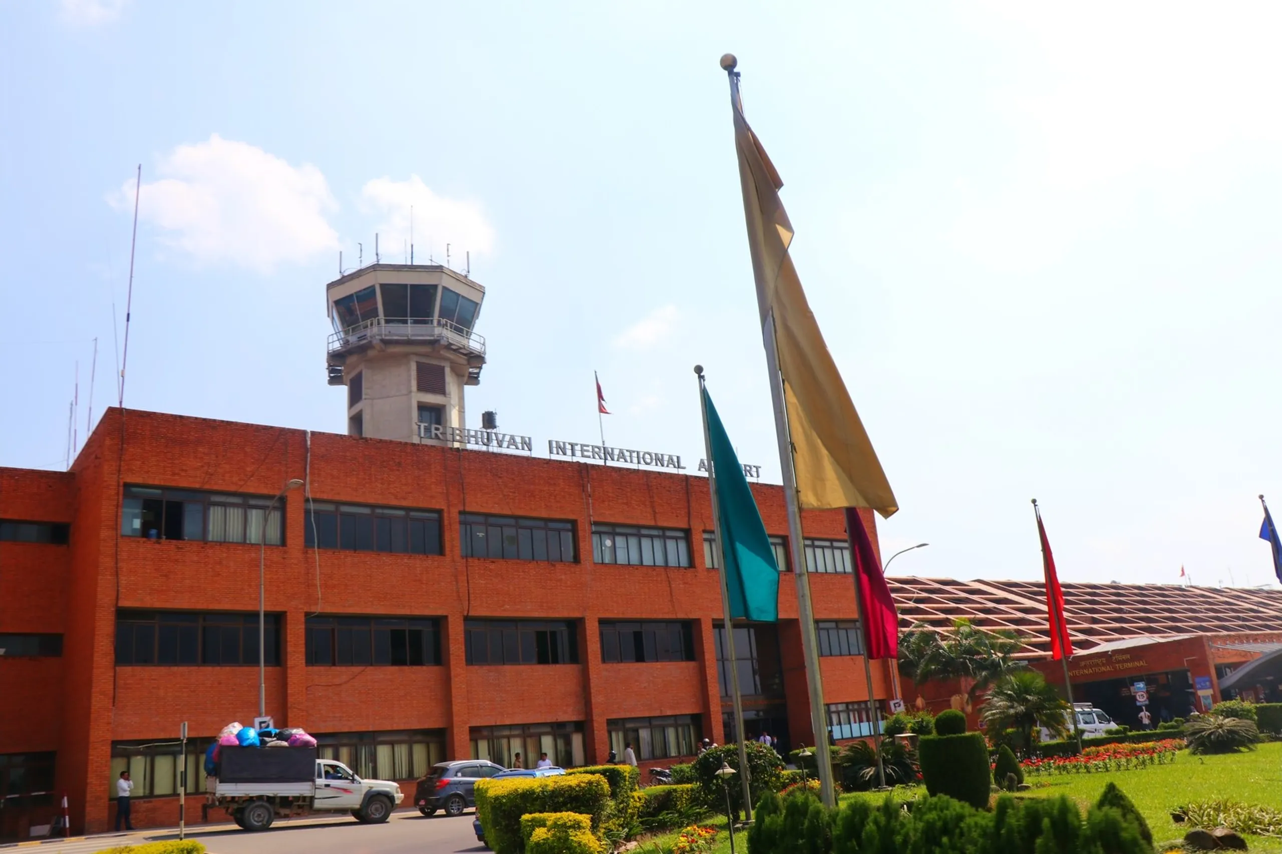 A clear bright exterior photograph of Tribhuvan International Airport in Kathmandu, Nepal - the iconic red-brick multi-story terminal building fills most of the frame with large white lettering spelling "TRIBHUVAN INTERNATIONAL AIRPORT" across the upper facade below an air traffic control tower flying a small Nepali flag, multiple colorful flags - teal, gold, red - mounted on tall poles in the foreground landscaped garden area with manicured hedges, flower beds and a palm tree, a white pickup truck with cargo visible parked to the far left, vehicles and people moving in front of the terminal building, a smaller "International Terminal" sign partially visible to the right, all under a bright blue sky with wispy clouds - the final scene marking the triumphant return of the entire Shey Phoksundo trekking group to the capital after one of Nepal's most remote and rewarding Himalayan adventures, Tribhuvan International Airport Kathmandu Nepal, TIA Kathmandu Nepal arrival, Nepal capital Kathmandu airport, Shey Phoksundo trek return Kathmandu, Nepal domestic international airport photography, end of trek Nepal Kathmandu homecoming