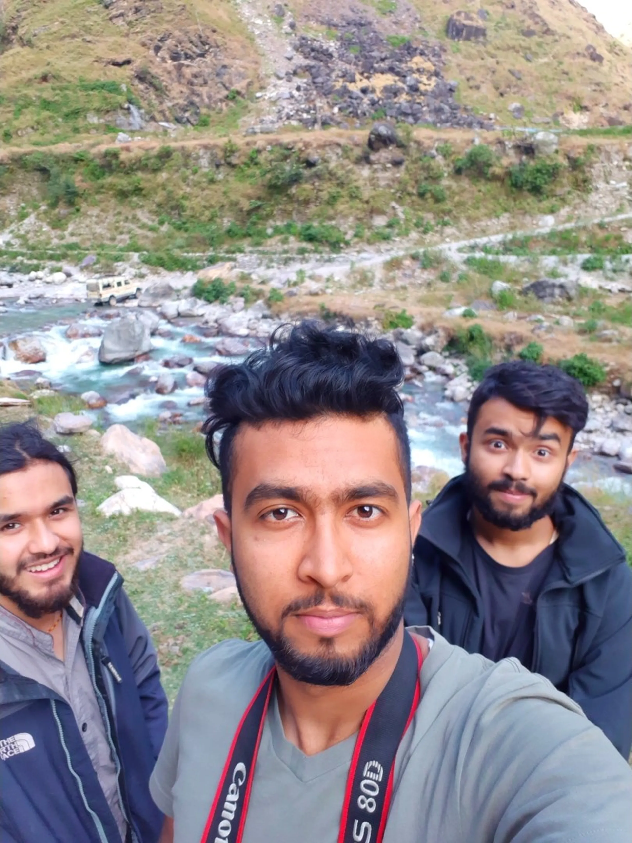 A selfie taken by the central trekker holding a Canon DSLR camera by its red-labeled strap, capturing three young South Asian male trekkers against a stunning Himalayan river gorge backdrop - a rushing turquoise glacial river winding through a rocky valley flanked by steep green hills, a white jeep visible on the distant trail above, all three men wearing layered trekking jackets (grey North Face, olive grey, and dark green), one making a wide-eyed funny expression, Canon camera strap visible, Nepal trekking selfie, Canon photography Nepal mountains, Shey Phoksundo trek friends, Himalayan river gorge adventure, batch mates Nepal trek