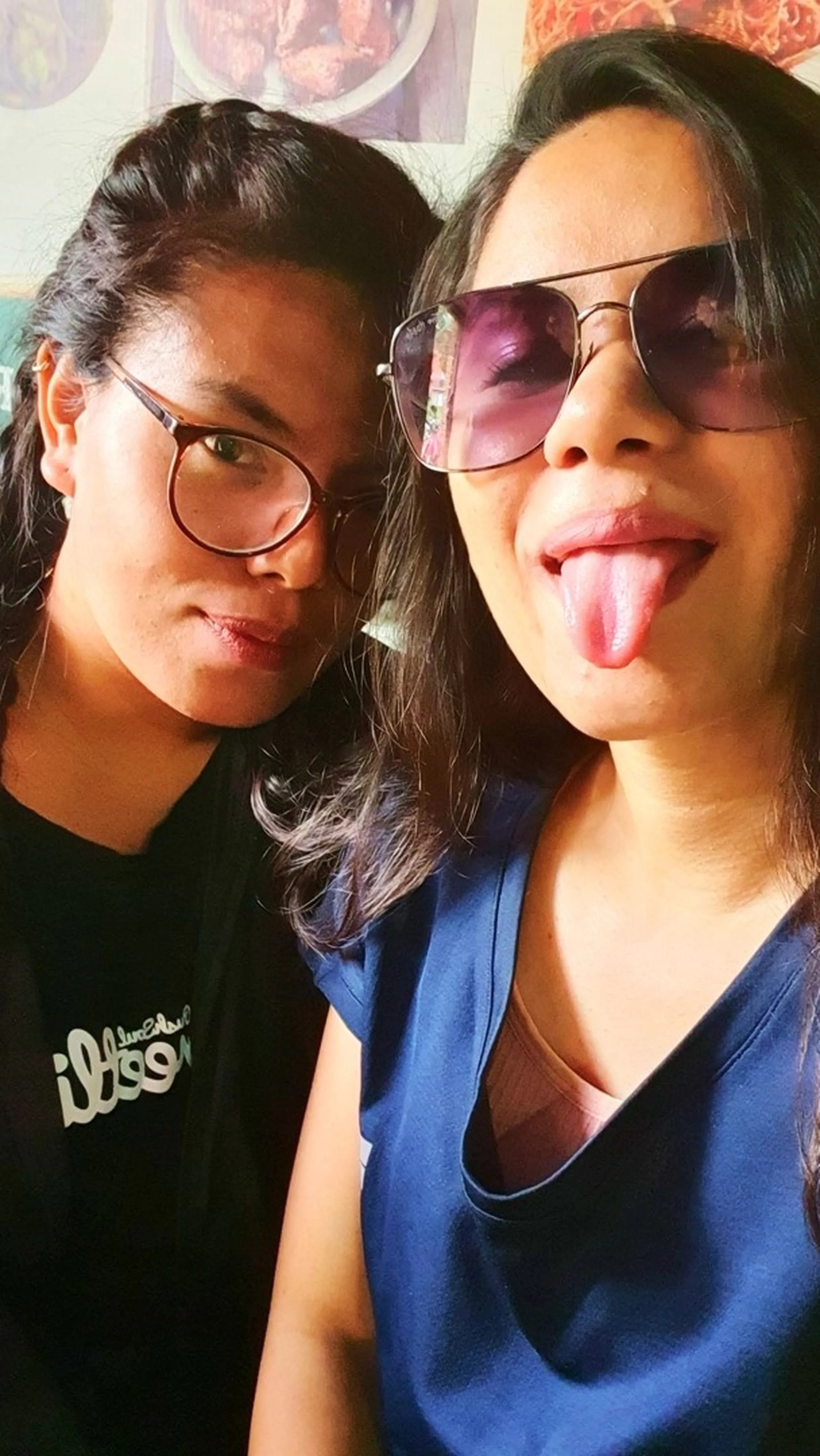 Two young Nepali women taking a fun indoor selfie before a hiking trip in Nepal - one wearing round glasses and a black t-shirt making a pouty face, the other in a blue top and large purple-tinted aviator sunglasses sticking her tongue out playfully; colorful food posters visible in the background, capturing the carefree pre-trek excitement of friends heading to Manikhel Lalitpur Nepal