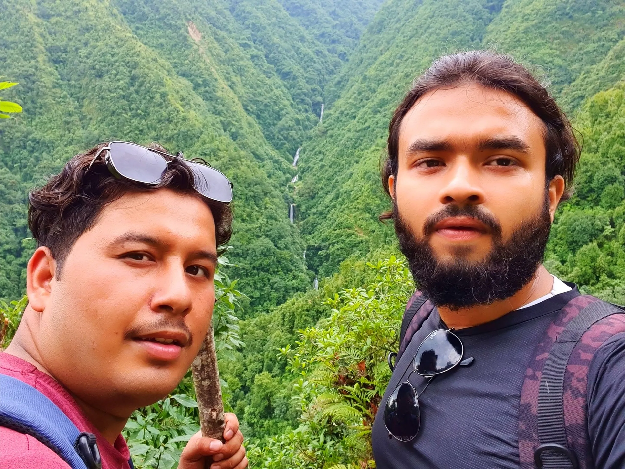 Two male trekkers taking a close-up selfie at the Simba Falls viewpoint in Manikhel Lalitpur Nepal - one clean-shaven with sunglasses on his head holding a trekking stick in a maroon shirt, the other bearded with sunglasses hanging from his collar wearing a dark patterned athletic top; in the background a spectacular deep green jungle gorge with multiple tiers of white Simba waterfall cascades framed between steep forested hills - epic Nepal waterfall trek friends travel photo