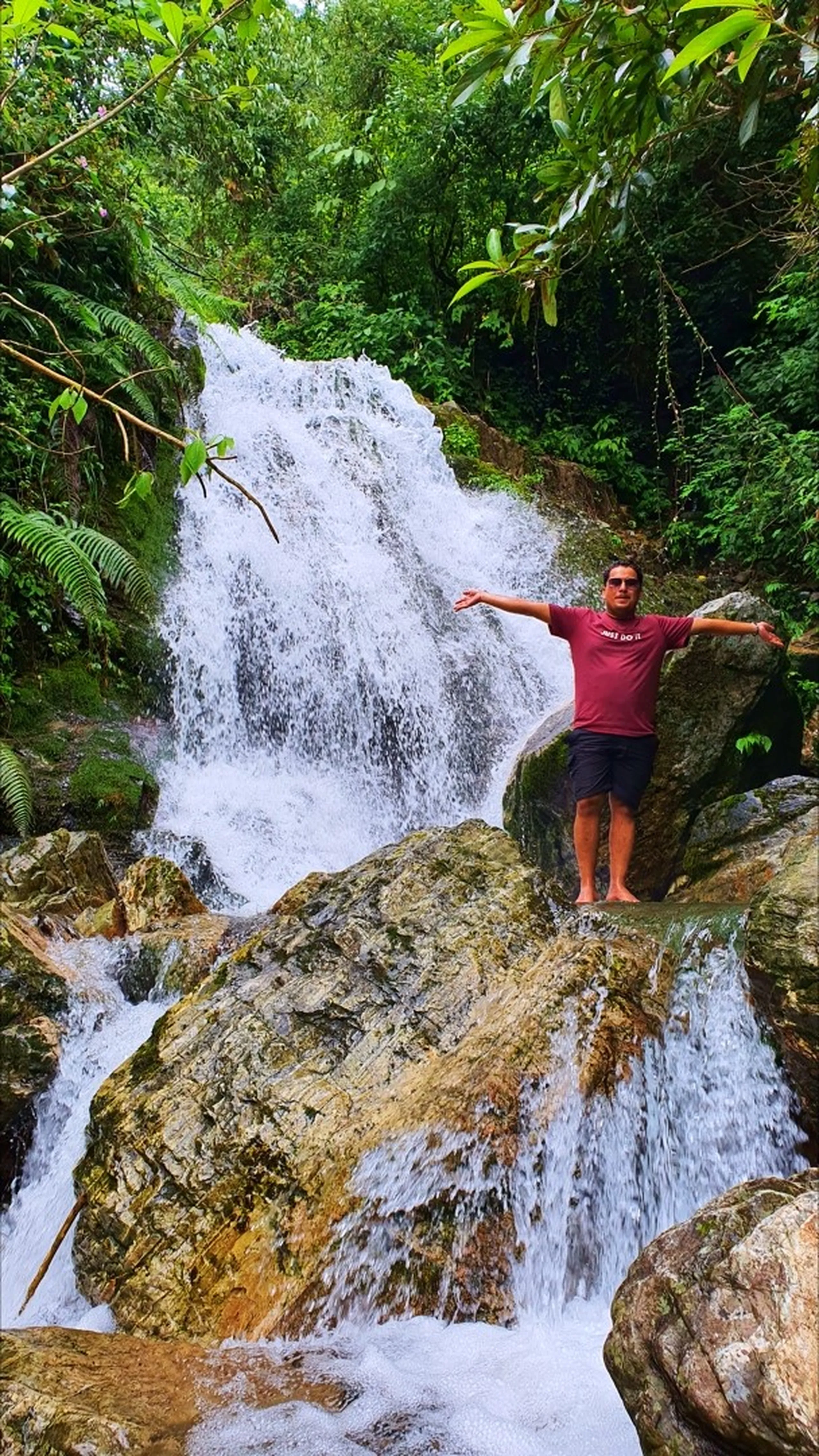 A male trekker in a maroon Nike t-shirt and dark shorts standing barefoot on a mossy boulder with arms outstretched wide in a triumphant pose in front of Simba Waterfall in Manikhel Lalitpur Nepal; powerful white water cascades down multi-level rocky tiers behind him, surrounded by tropical green jungle with ferns and broad-leafed trees - joyful achievement moment at Simba Falls Nepal hidden waterfall near Kathmandu Valley trekking