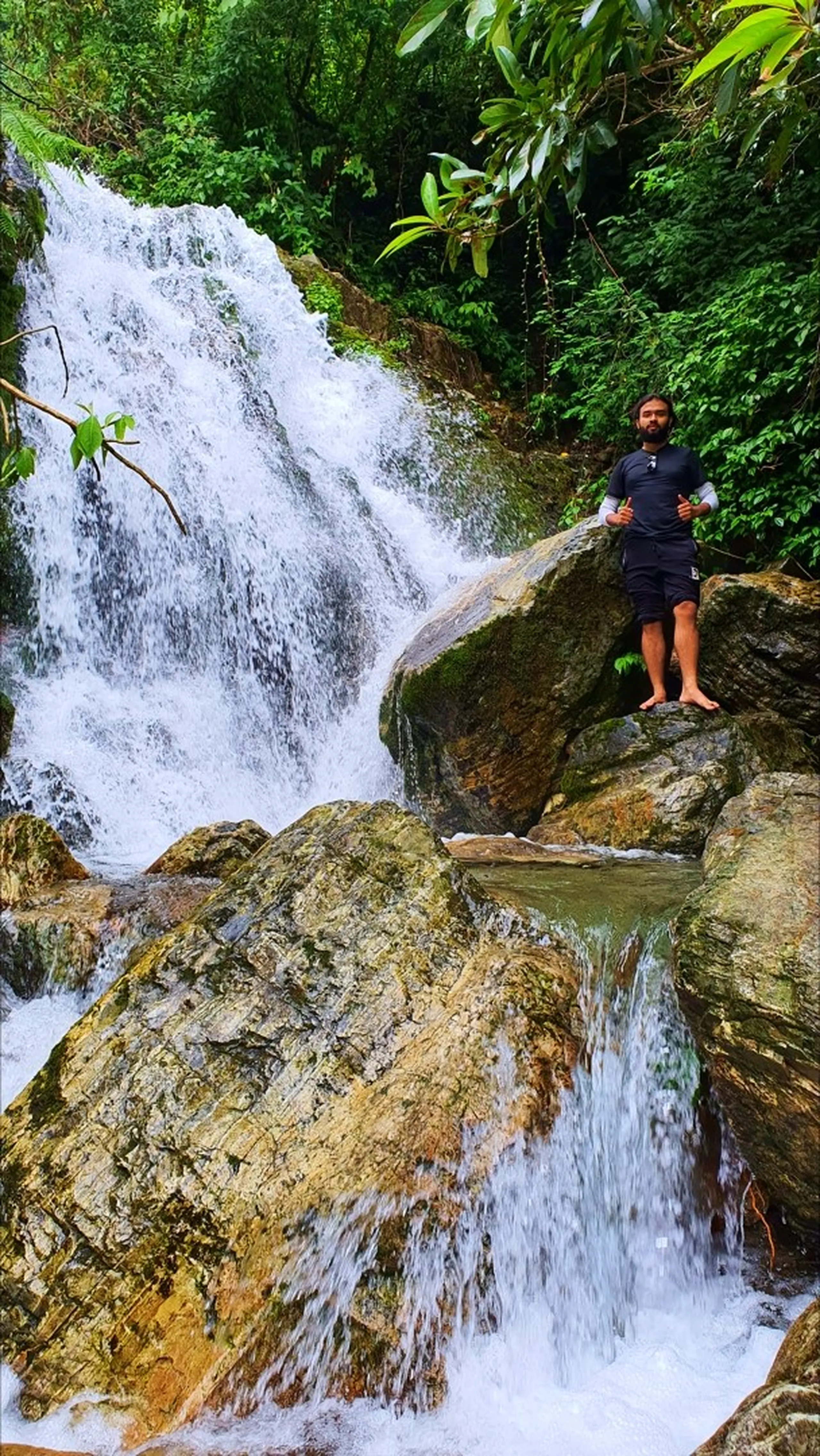 A bearded male trekker in a black rash guard over white long sleeves and dark shorts standing barefoot giving a thumbs-up on a moss-covered boulder beside Simba Falls in Manikhel Lalitpur Nepal; strong white waterfall cascading over multi-step rocky ledges on the left, golden-hued mossy rocks in the middle ground, green tropical jungle canopy overhead - adventure trekking Nepal Simba Waterfall Lalitpur near Kathmandu