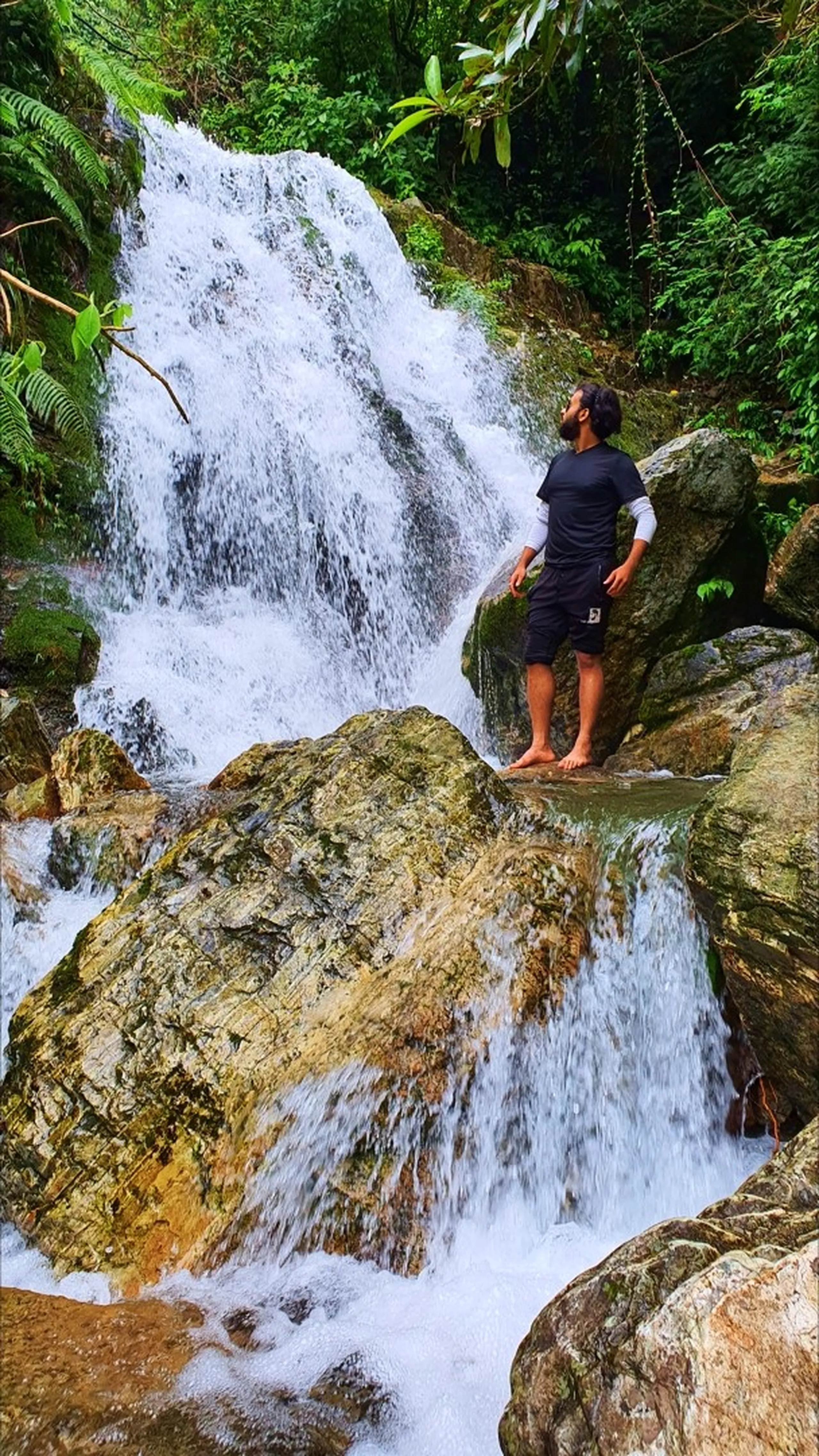 A bearded male trekker in a black and white athletic outfit standing barefoot on a mossy boulder looking upward contemplatively at Simba Waterfall in Manikhel Lalitpur Nepal; powerful white multi-tiered waterfall cascading down dark rocks behind him, moss-covered golden boulders with rushing water in the foreground, lush tropical ferns and green jungle surrounding - peaceful Nepal waterfall moment, Simba Falls Lalitpur off-beat trek near Kathmandu