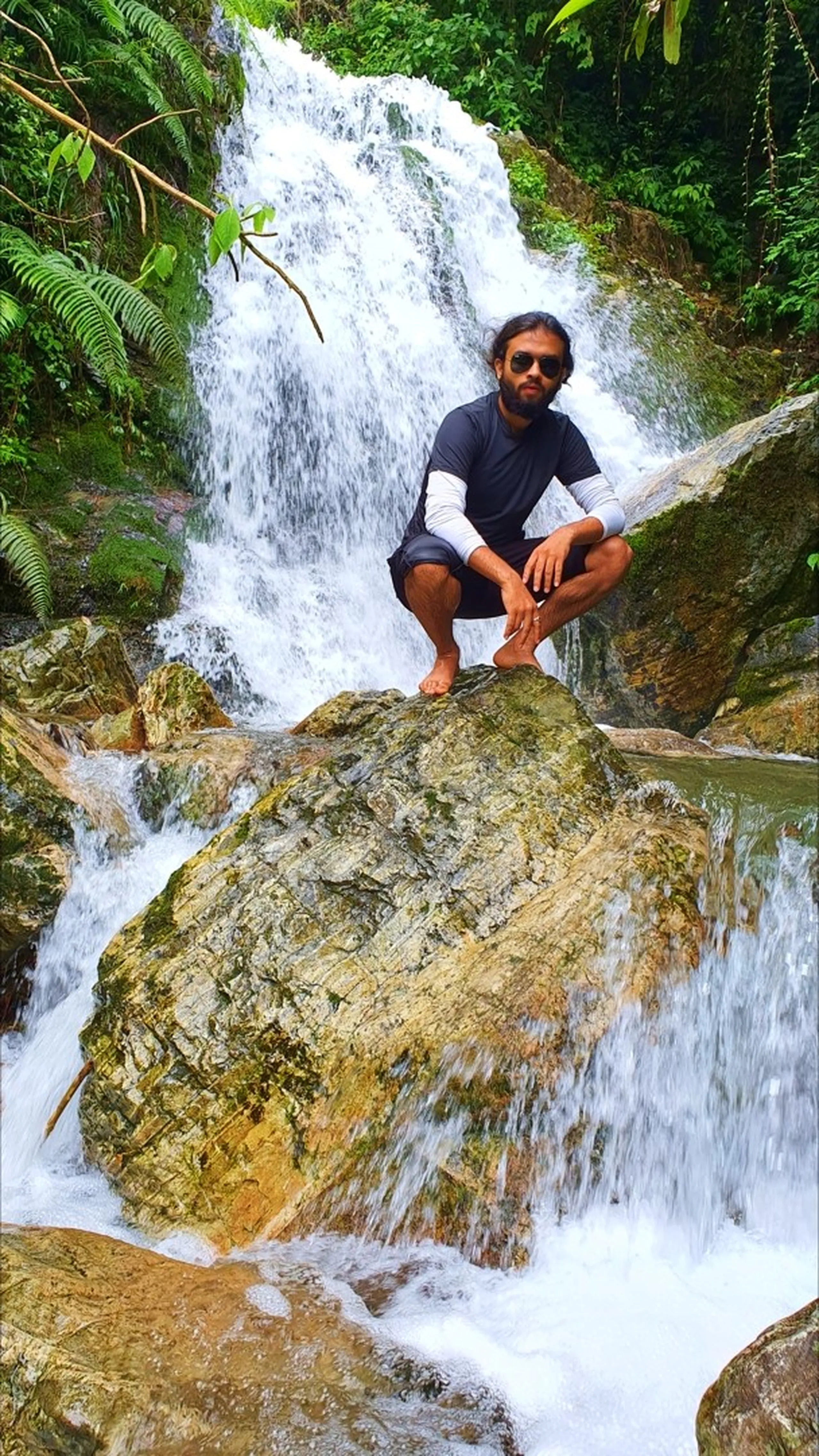 A bearded male trekker in a black rash guard layered over white long sleeves and dark shorts squatting confidently on a large golden moss-covered boulder at the base of Simba Waterfall in Manikhel Lalitpur Nepal; wearing aviator sunglasses and barefoot, white cascading water rushing around the boulder on both sides, dense green jungle with ferns and tropical trees framing the entire scene - cool Nepal waterfall pose, Simba Falls Lalitpur hidden gem trek near Kathmandu Valley