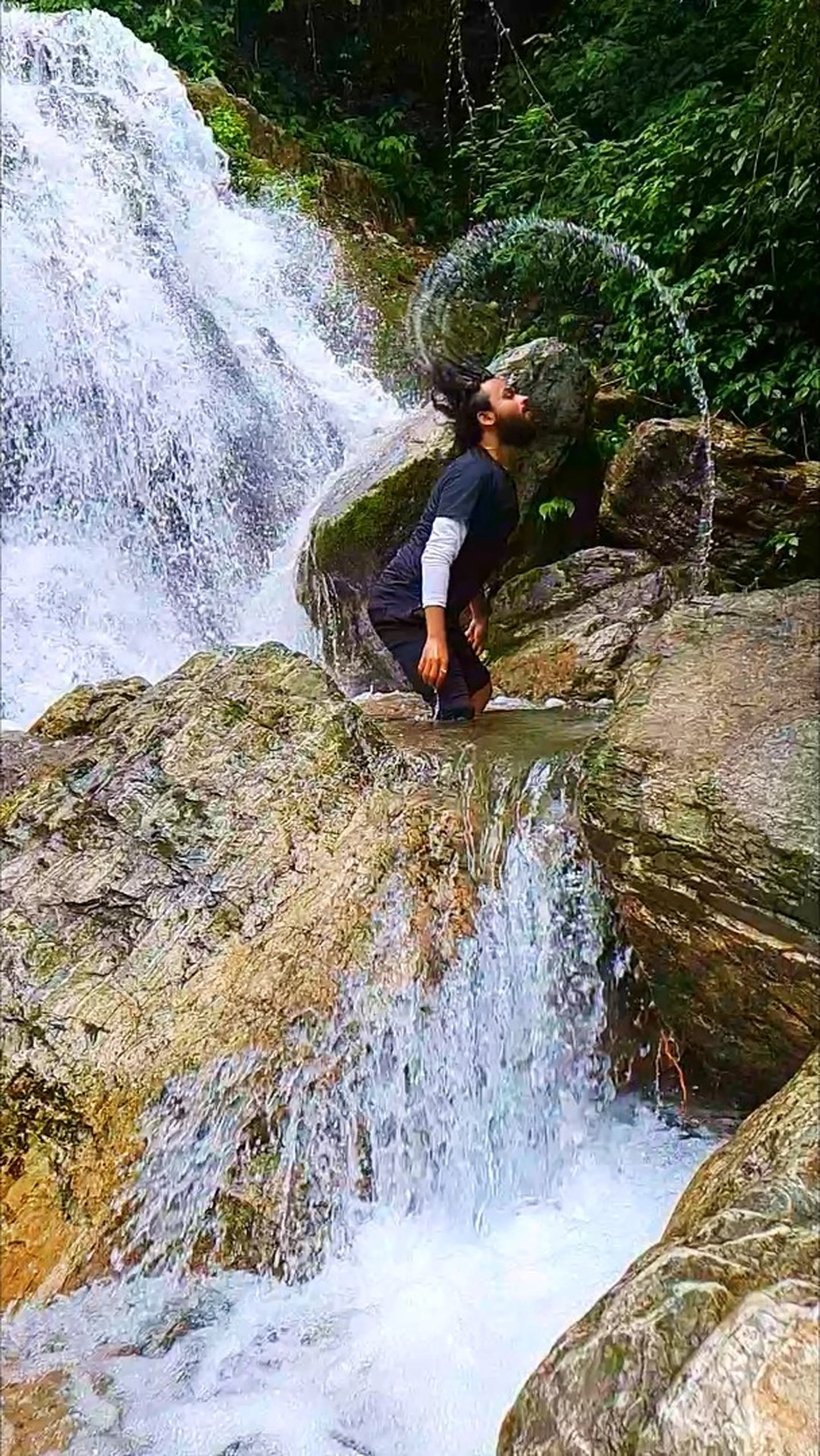 A bearded male trekker in a black and white long-sleeve athletic shirt caught mid-hair-flip at Simba Falls in Manikhel Lalitpur Nepal - standing waist-deep in a natural rock pool between two rushing white water cascades, head thrown back dramatically with wet hair arcing through the air creating a water spray halo; golden moss-covered boulders surrounding him, dense green jungle in the background - wild and free Nepal waterfall moment, Simba Fall Lalitpur hidden waterfall adventure near Kathmandu