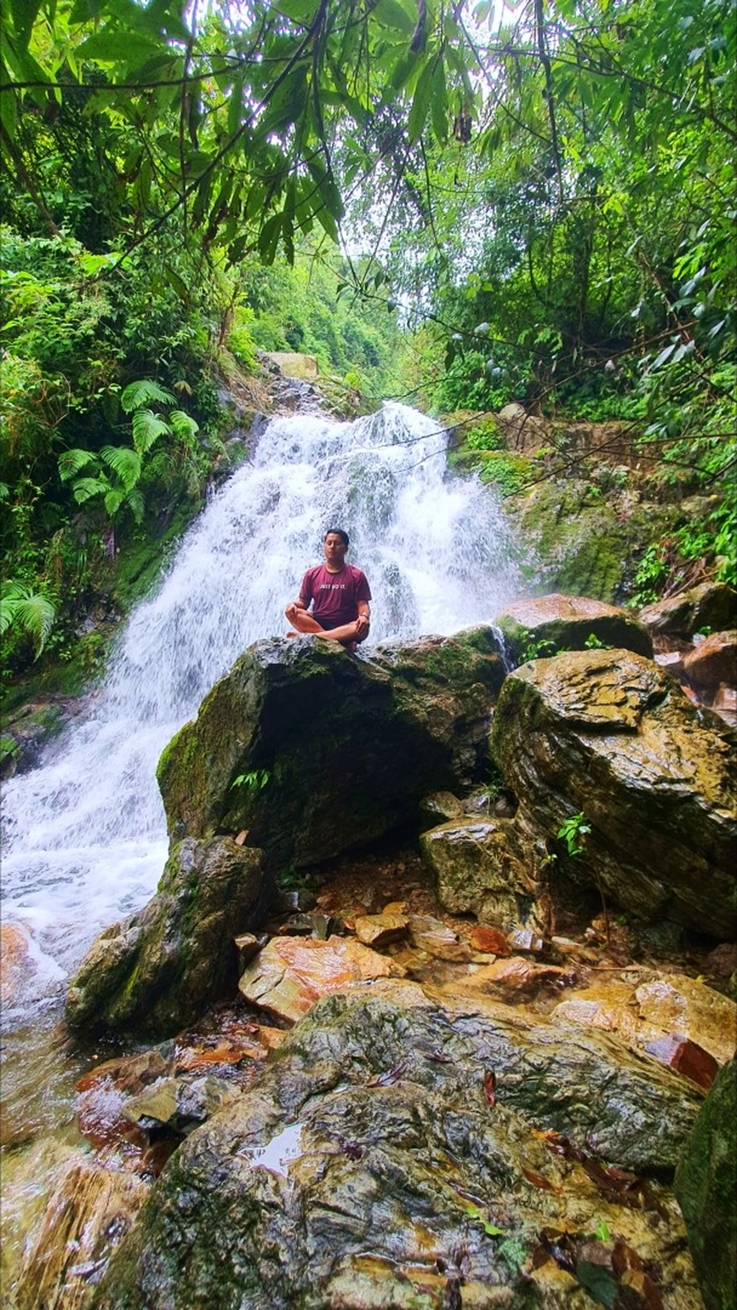 A male trekker in a maroon Nike 'Just Do It' t-shirt sitting in a perfect cross-legged lotus meditation pose on a large flat moss-covered boulder directly in front of Simba Waterfall in Manikhel Lalitpur Nepal; powerful white cascading water rushes down layered rocky tiers behind him, surrounded by a cathedral of lush tropical ferns, overhanging tree branches, and dense monsoon jungle - serene waterfall meditation Nepal, Simba Falls Lalitpur mindfulness trekking near Kathmandu Valley