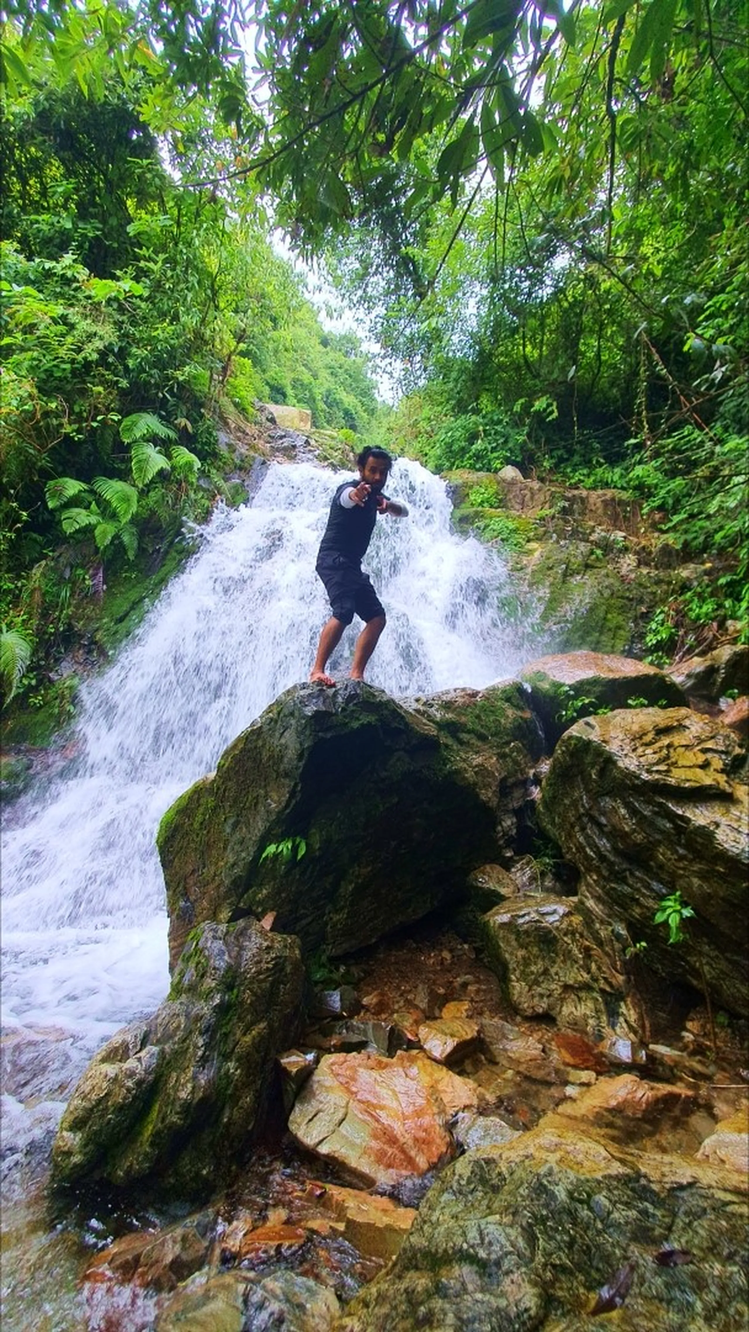 A male trekker in all-black shorts and t-shirt standing barefoot on a large moss-covered boulder at Simba Falls Manikhel Lalitpur Nepal, pointing finger-guns playfully at the camera with a confident grin; powerful white multi-tier waterfall cascading down behind him, golden and rust-colored rocky riverbed in the foreground, tropical ferns and lush green jungle canopy overhead - fun Nepal waterfall trekking moment, Simba Fall Lalitpur adventure destination near Kathmandu