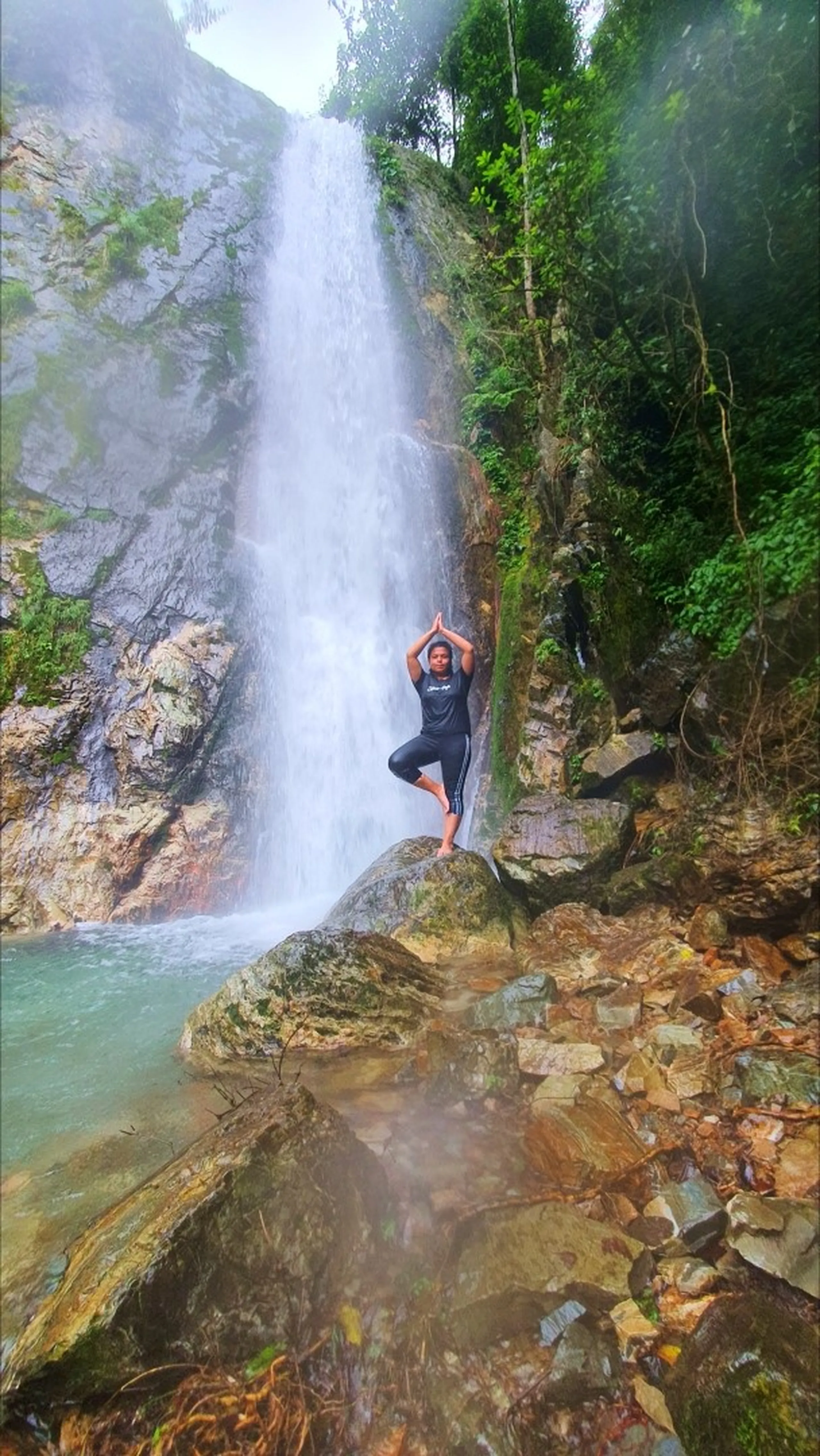 A woman in all-black athletic wear performing a graceful tree pose yoga stance barefoot on a mossy boulder directly in front of the towering main drop of Simba Falls in Manikhel Lalitpur Nepal - standing on one leg with hands pressed together overhead in namaskar mudra; the tall narrow waterfall plunges dramatically behind her down a sheer grey cliff face into a turquoise natural pool, jungle greenery framing both sides - Nepal waterfall yoga pose, Simba Falls Lalitpur wellness trekking hidden gem near Kathmandu