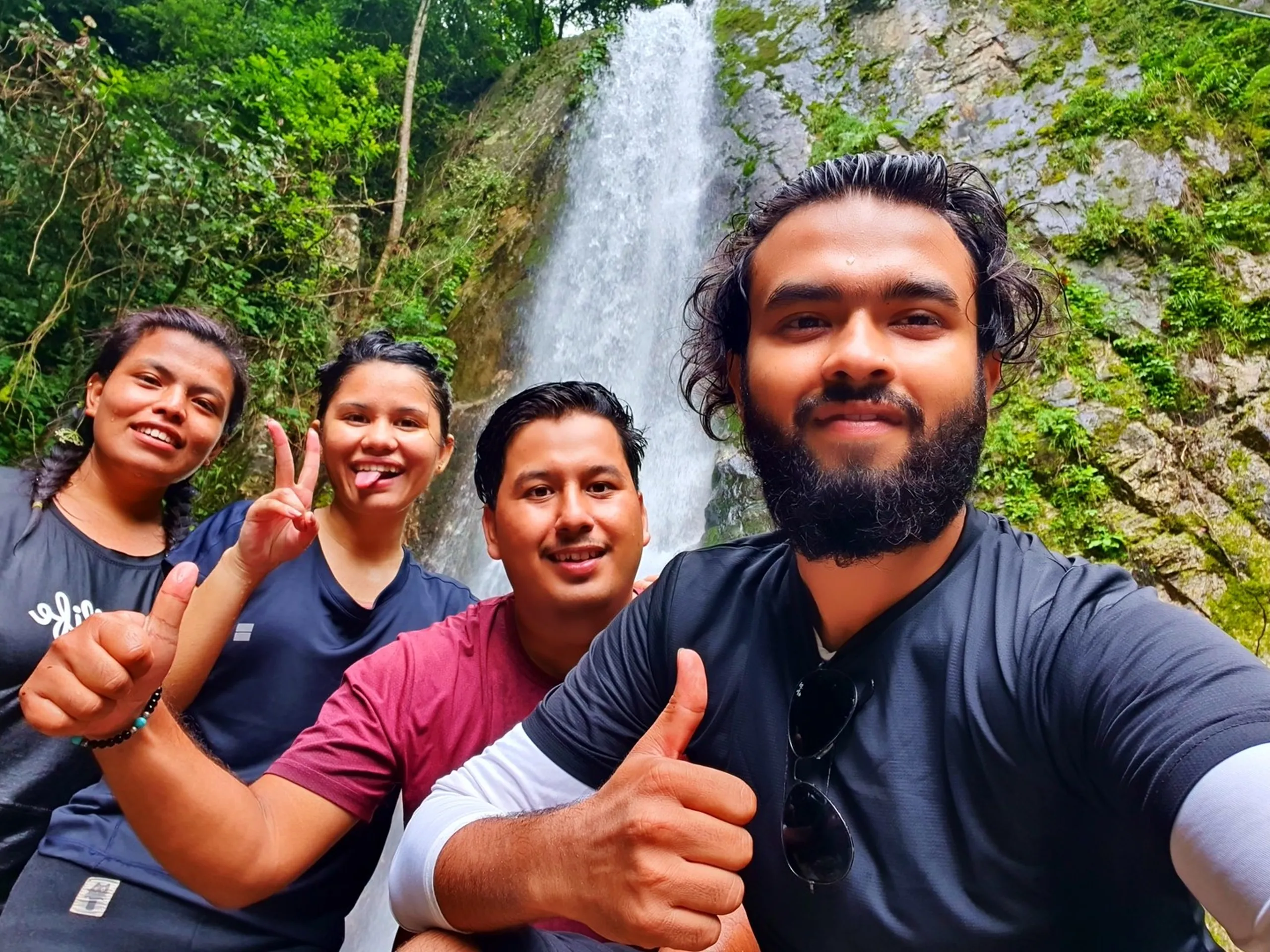 A cheerful group of four Nepali friends - two women and two men - taking a selfie with the towering main drop of Simba Falls in Manikhel Lalitpur Nepal prominently visible behind them; one woman gives a thumbs-up and tongue-out, another flashes a peace sign, a man in maroon smiles broadly, and the bearded man taking the selfie gives a big thumbs-up; all appear wet and exhilarated - happy group trekking Nepal waterfall selfie, Simba Fall Lalitpur friends adventure near Kathmandu