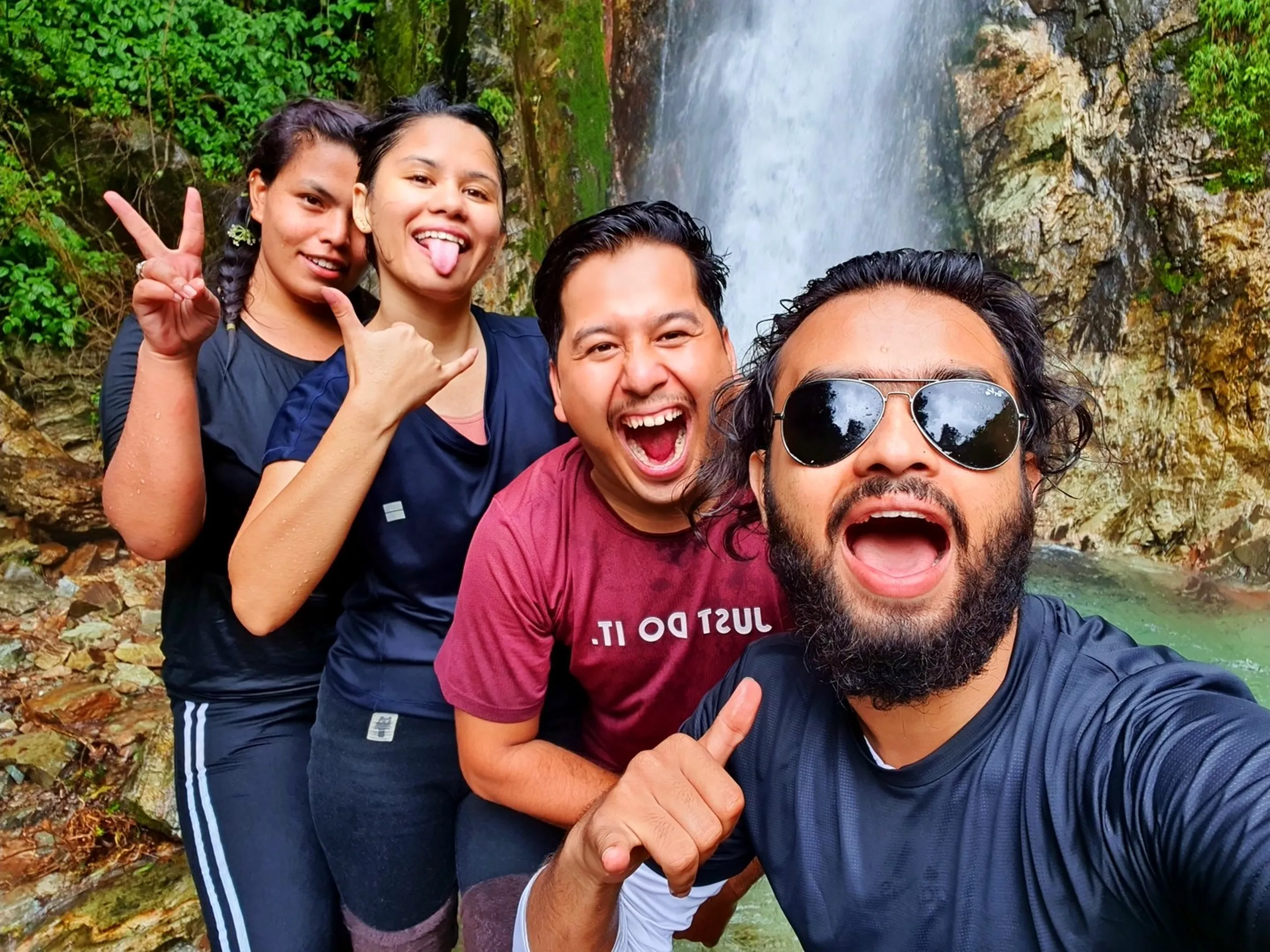 Four drenched and ecstatic Nepali trekkers taking a goofy wide-mouthed selfie at Simba Falls Manikhel Lalitpur Nepal - all soaking wet with big open-mouthed laughing expressions; one woman flashes a peace sign, a man in maroon Nike shirt screams with joy, and the bearded selfie-taker in a dark shirt with aviator sunglasses shouts in delight with a thumbs-up; mossy rocks and a turquoise waterfall pool visible behind them - pure joy Nepal waterfall fun, Simba Fall Lalitpur group trekking adventure near Kathmandu