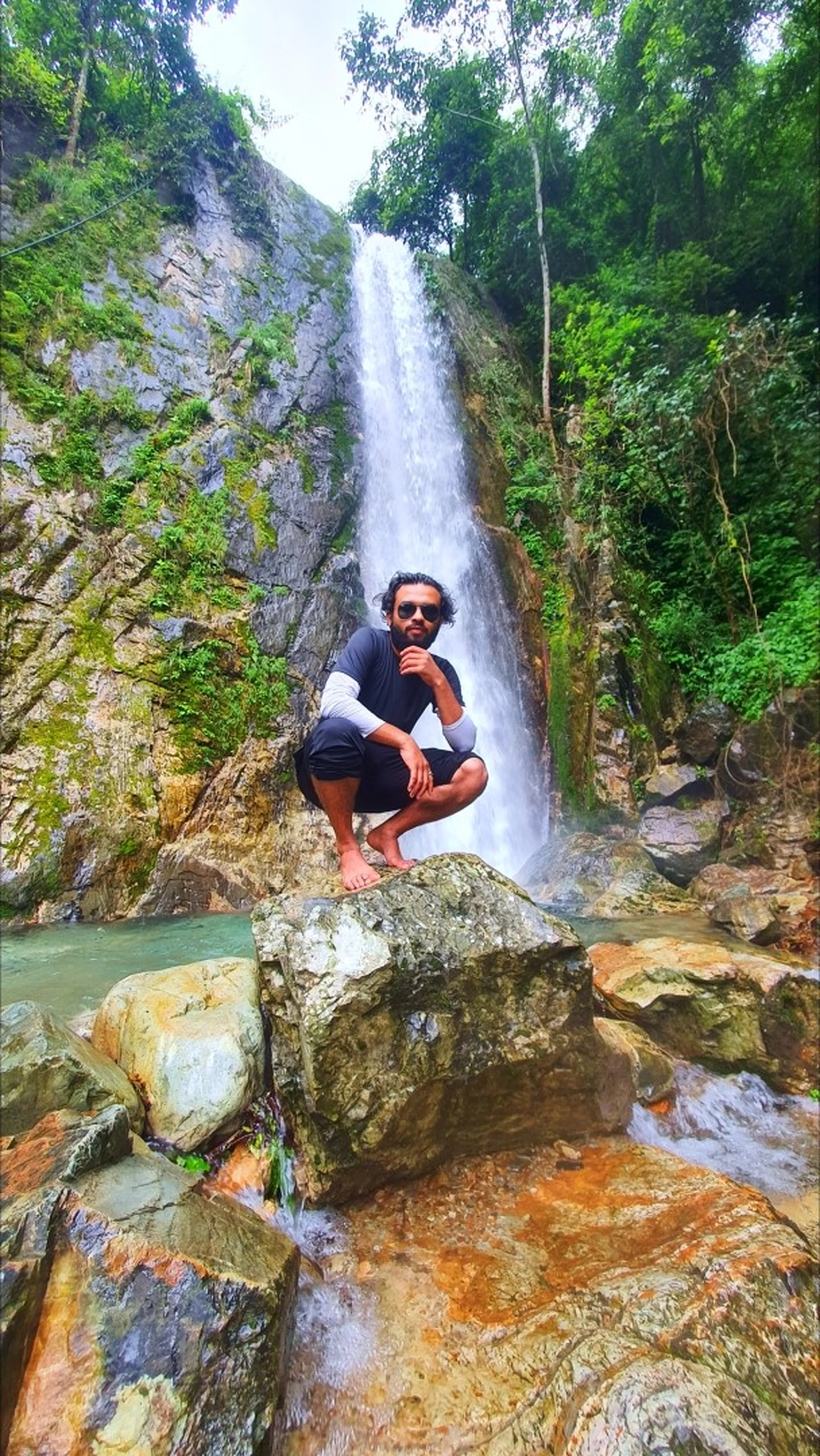 A bearded male trekker in a white and black long-sleeve athletic outfit crouching on a large boulder in a cool posed squat at the base of the main single-drop Simba Falls in Manikhel Lalitpur Nepal - the tall narrow waterfall descends a sheer pale grey cliff face directly behind him into a clear turquoise pool, with green moss and jungle vegetation clinging to the rock walls on both sides; water flowing around the base rocks - stylish Nepal waterfall portrait, Simba Falls Lalitpur tall waterfall near Kathmandu trekking