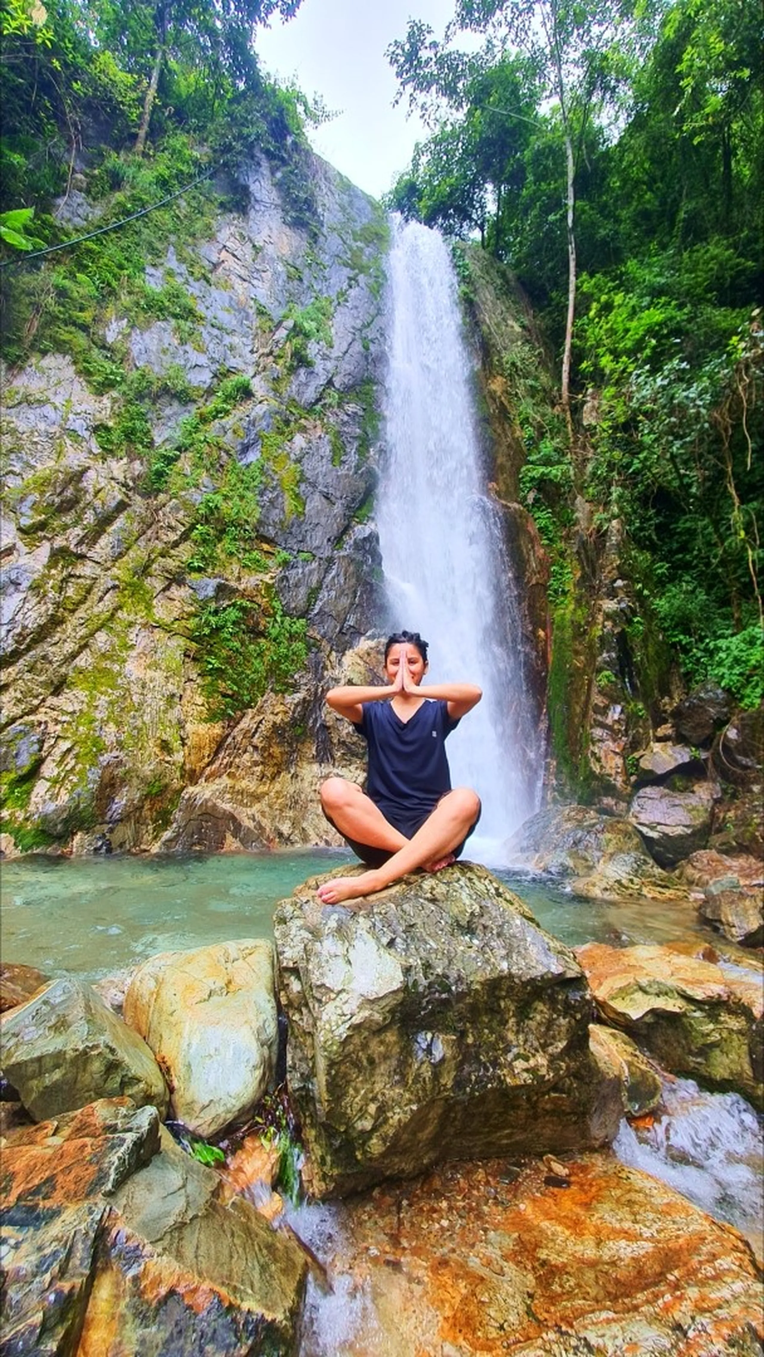 A young woman in a black outfit performing a cross-legged seated meditation pose with hands in namaste prayer at chest level on a flat boulder in the natural pool at the base of the towering Simba Falls in Manikhel Lalitpur Nepal; the sheer single-drop waterfall crashes down a tall grey cliff directly behind her into a clear green-blue plunge pool, mossy jungle-covered rock walls on both sides, overcast sky overhead - peaceful Nepal waterfall meditation yoga, Simba Fall Lalitpur spiritual trekking destination near Kathmandu