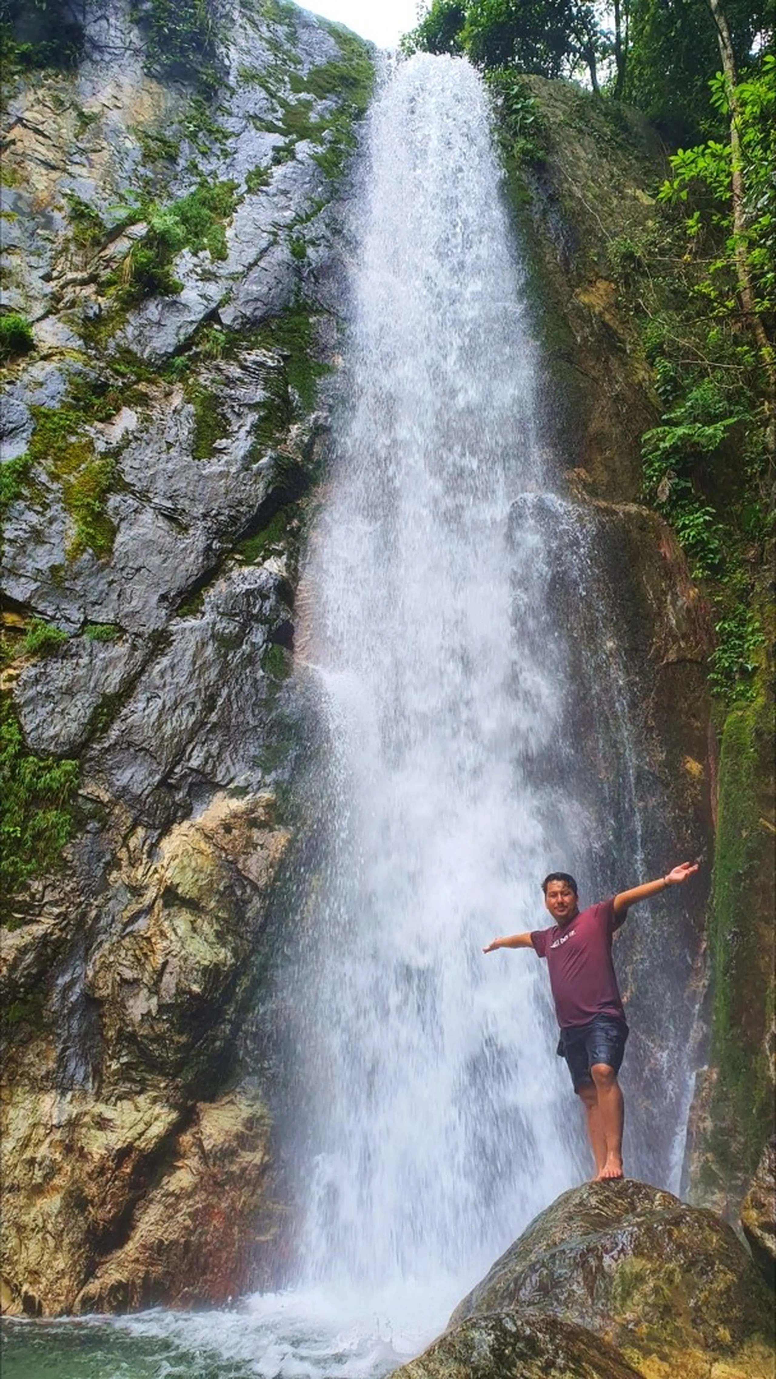 A male trekker in a maroon t-shirt and dark shorts standing barefoot on a large boulder with arms spread wide in complete freedom and exhilaration directly beneath the towering main drop of Simba Falls in Manikhel Lalitpur Nepal; the powerful single-stream waterfall plunges the full height of a sheer grey rock face behind him, waterfall mist rising around his feet, jungle trees and green shrubs clinging to the cliff edges - triumphant Nepal waterfall moment, Simba Falls Lalitpur tall waterfall adventure near Kathmandu