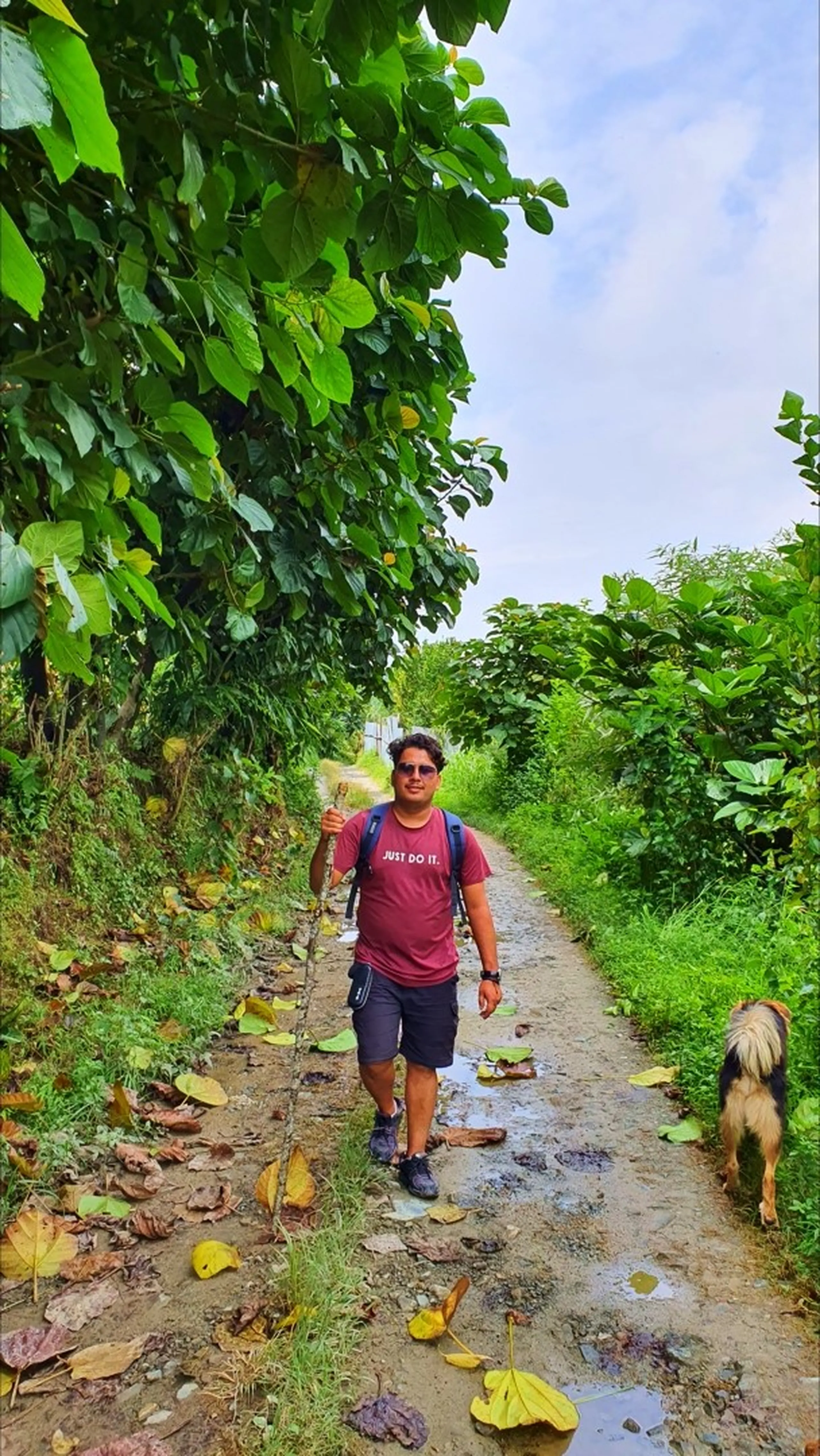 A male hiker in a maroon Nike 'Just Do It' t-shirt, dark shorts, and sunglasses walking confidently down a muddy narrow village trail flanked by enormous tropical teak-like trees with oversized leaves in Manikhel Lalitpur Nepal; fallen yellow leaves scattered on the wet dirt path, a friendly local dog trotting alongside, monsoon sky overhead - classic Nepal village hiking trail experience near Kathmandu