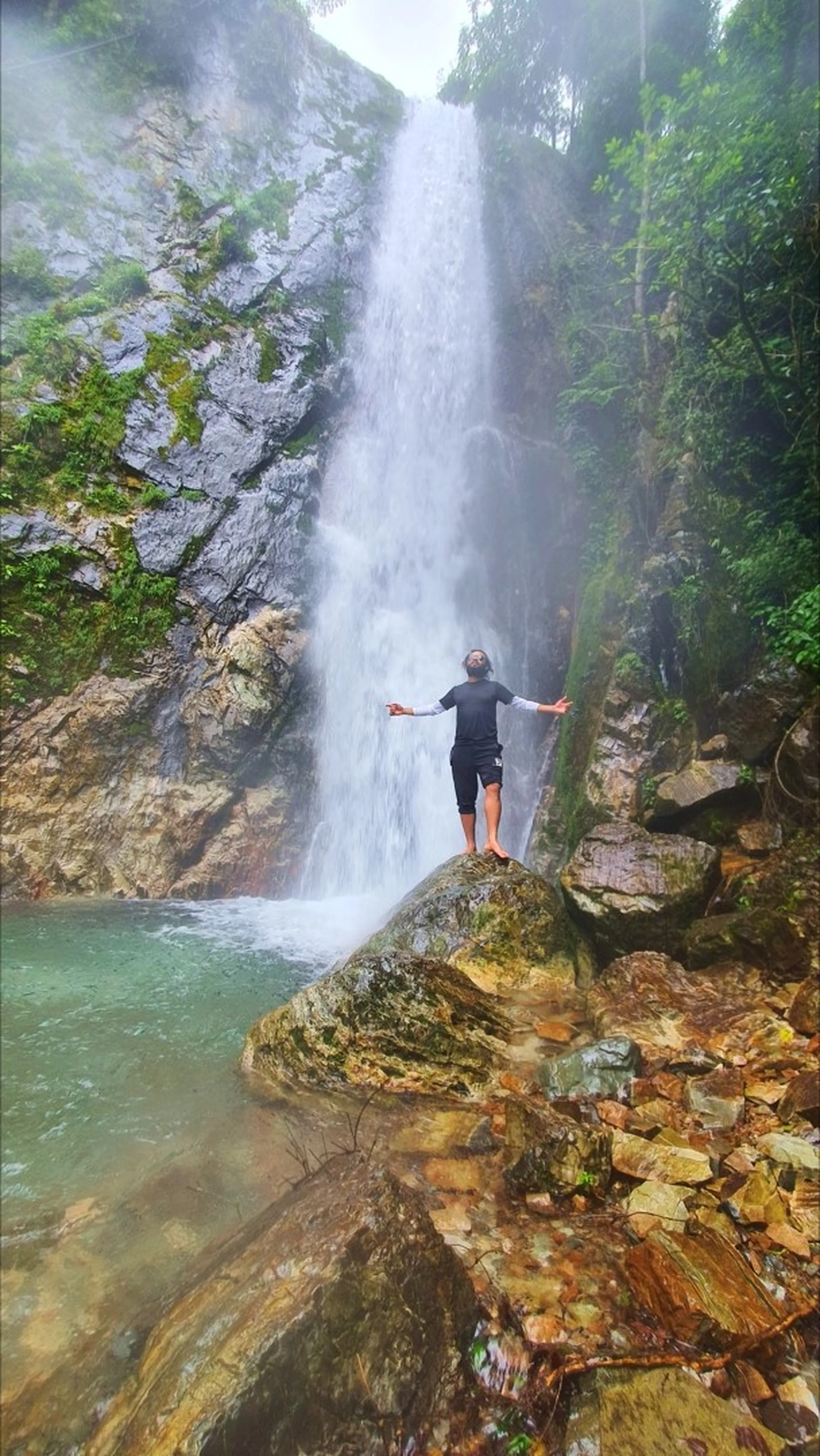 A male trekker in a black athletic shirt and shorts standing barefoot on a large boulder with arms outstretched wide, face tilted upward toward the sky in an expression of complete surrender and bliss, directly beneath the tall thundering Simba Falls in Manikhel Lalitpur Nepal; the powerful waterfall crashes down a towering grey cliff face behind him generating fine mist, a clear turquoise natural pool at the base, jungle vegetation covering the surrounding rocky walls - spiritual Nepal waterfall freedom pose, Simba Fall Lalitpur monsoon waterfall near Kathmandu