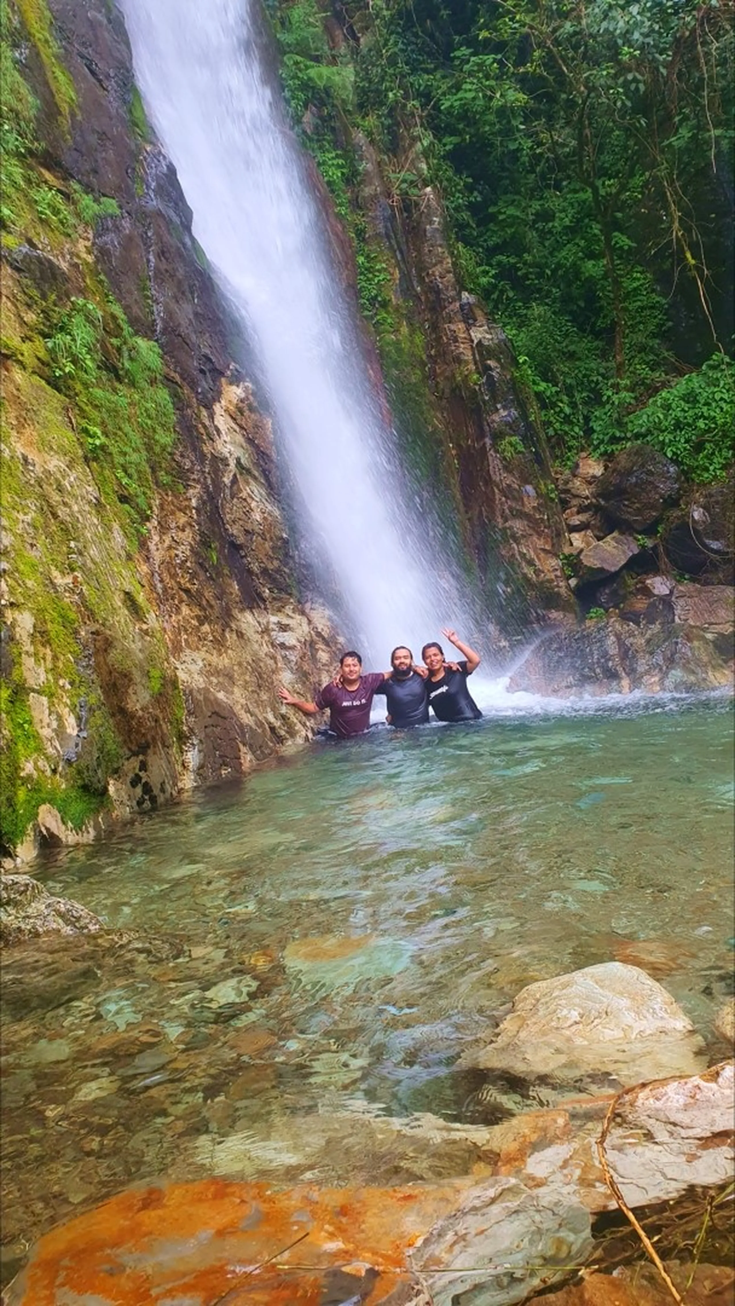 Three male trekkers standing waist-deep in the clear turquoise natural plunge pool directly at the base of the tall single-drop Simba Falls in Manikhel Lalitpur Nepal, laughing and raising their hands in celebration; the towering waterfall crashes powerfully behind them down a sheer moss-streaked rock cliff, mist rising around them, green jungle-covered walls rising on both sides - exhilarating Nepal waterfall swimming natural pool, Simba Falls Lalitpur plunge pool adventure near Kathmandu