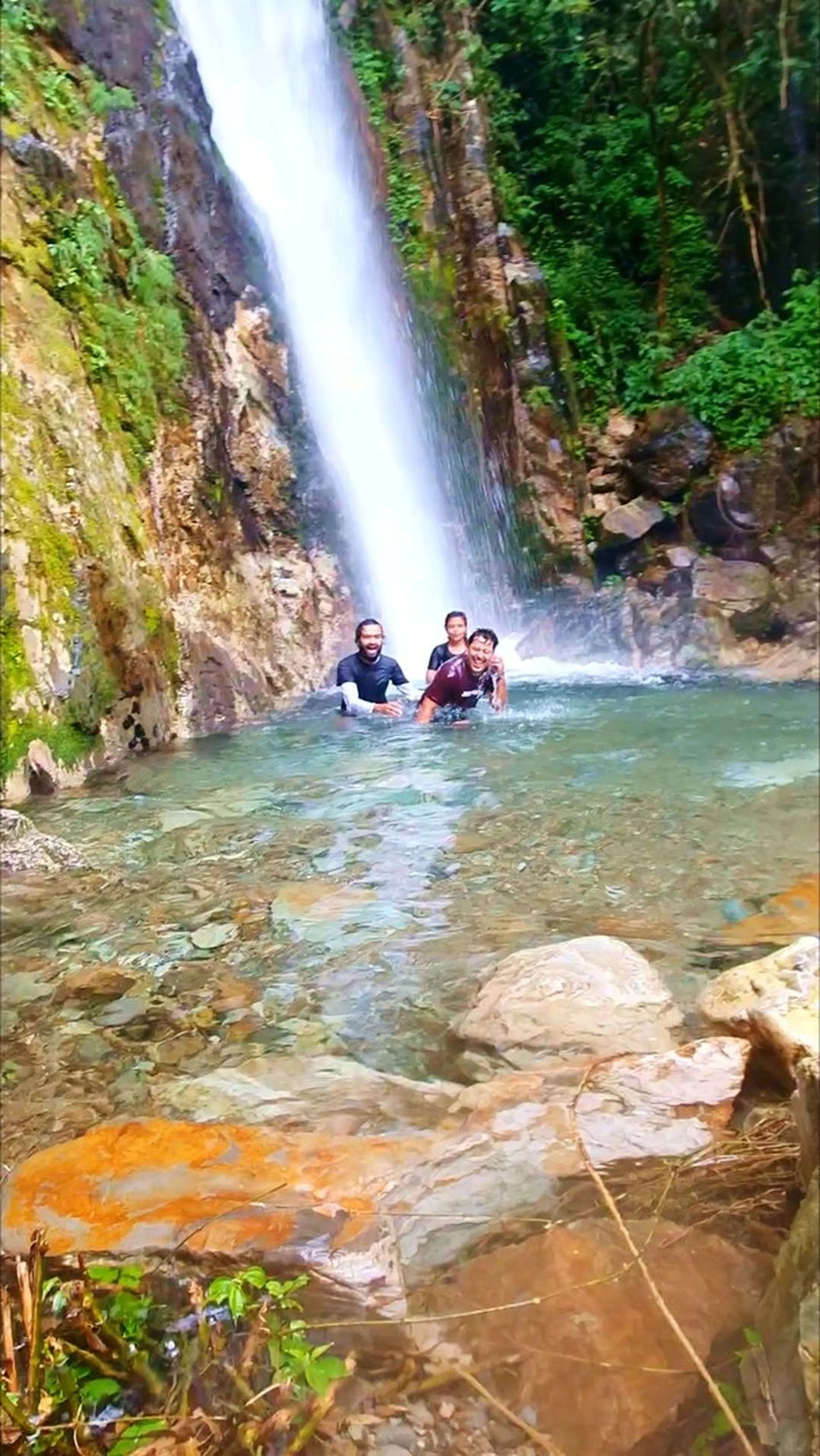 Two trekkers swimming and smiling in the crystal-clear turquoise natural pool at the base of Simba Falls in Manikhel Lalitpur Nepal - a bearded man in a black rash guard and a woman in a maroon outfit, both waist-deep in the pool with the thundering waterfall plunging directly behind them; mossy cliff walls on either side, green jungle trees above the waterfall crest - refreshing Nepal waterfall swimming natural pool, Simba Fall Lalitpur natural swimming hole near Kathmandu trekking
