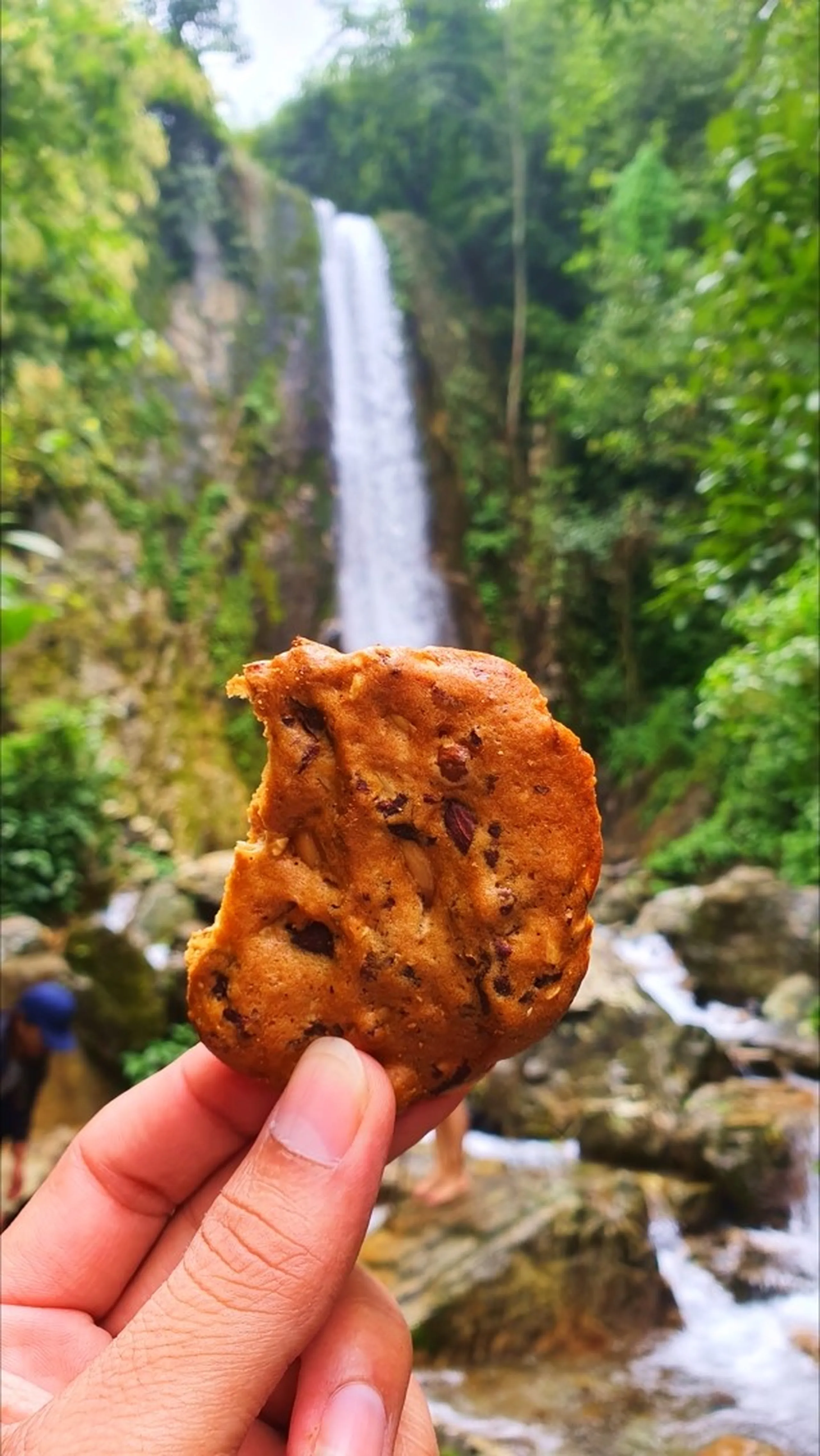 A close-up hand holding a half-eaten chocolate chip cookie in the foreground with the full-height Simba Falls in Manikhel Lalitpur Nepal dramatically blurred in the background - the tall slender waterfall drops down a sheer green jungle cliff face into rocky pools below, with dense tropical foliage on both sides and other trekkers faintly visible near the base; a playful trekking snack moment - Nepal waterfall hiking snack break, Simba Falls Lalitpur trekking food moment near Kathmandu Valley adventure trail