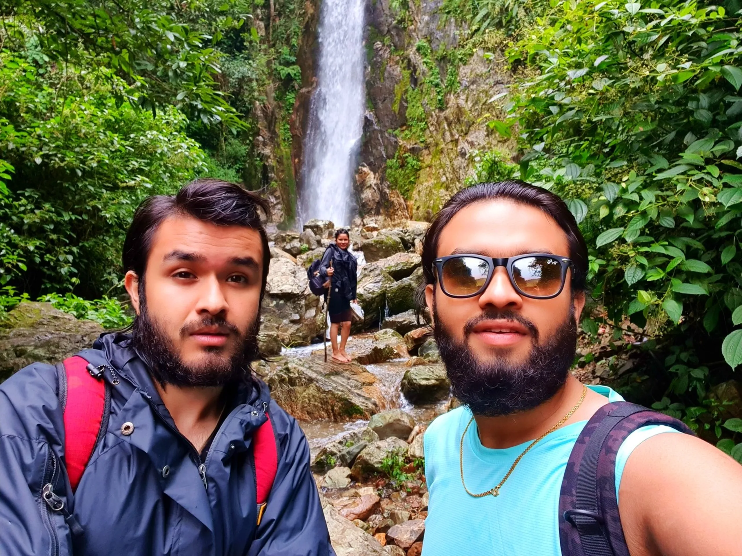 Two bearded male trekkers taking a close-up selfie at the base of Simba Falls in Manikhel Lalitpur Nepal - one in a dark navy jacket with a red backpack looking surprised, the other in a teal sleeveless top with brown-tinted wayfarers and a gold chain smiling confidently; in the soft-focus background a smiling woman in a black raincoat stands barefoot on the rocky riverbed with the towering waterfall cascading behind her, lush green jungle surrounding the entire scene - Nepal waterfall friends group selfie, Simba Fall Lalitpur day hike near Kathmandu