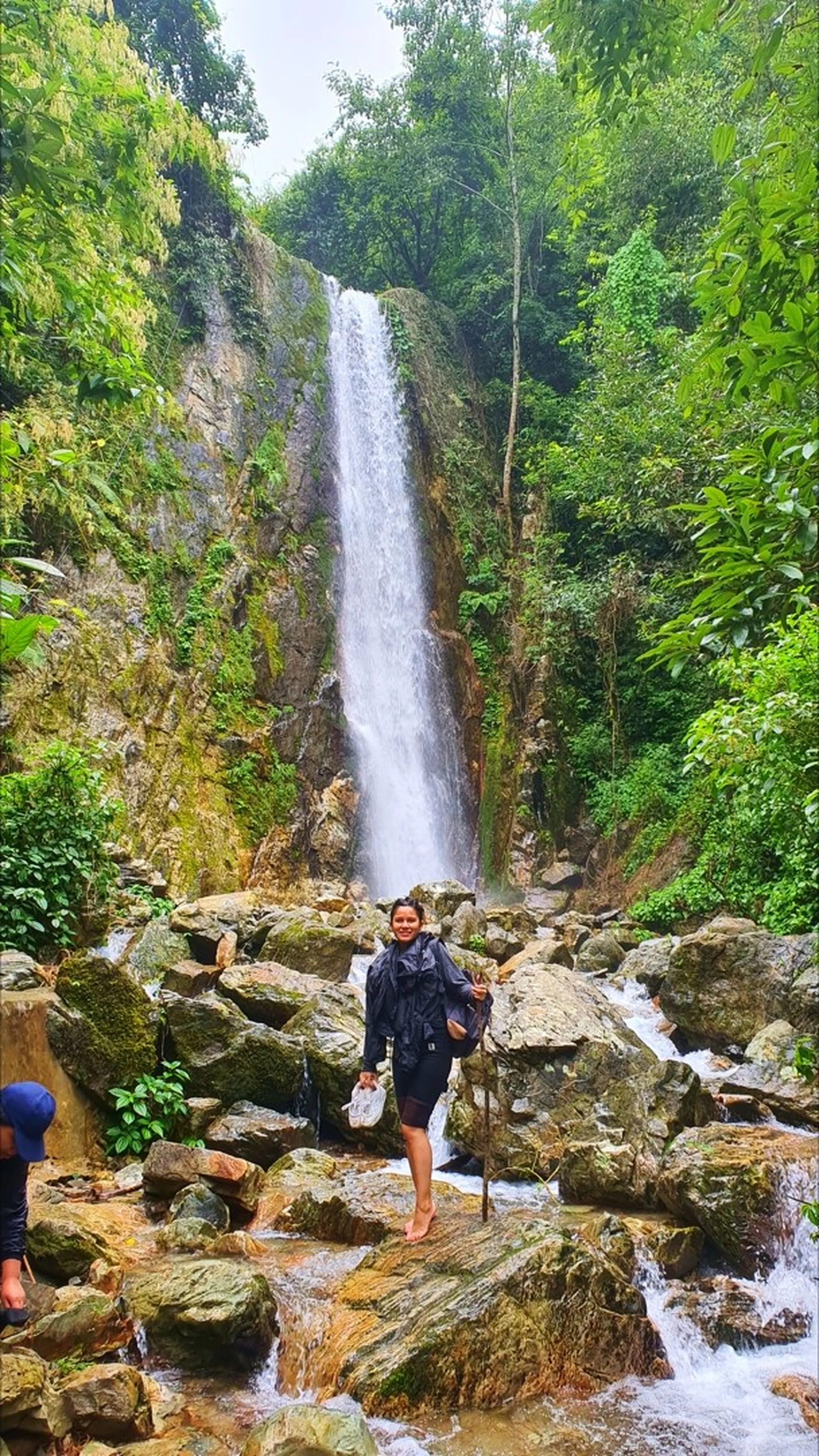 A smiling woman in a dark navy raincoat and black shorts standing barefoot and confidently on the wet mossy boulders at the base of the towering Simba Falls in Manikhel Lalitpur Nepal; the tall narrow single-drop waterfall cascades powerfully down a sheer grey rock cliff directly behind her, rushing water streaming over the rocky riverbed around her feet, dense green jungle vegetation on both sides - Nepal waterfall solo portrait female trekker, Simba Falls Lalitpur beautiful waterfall near Kathmandu adventure trekking