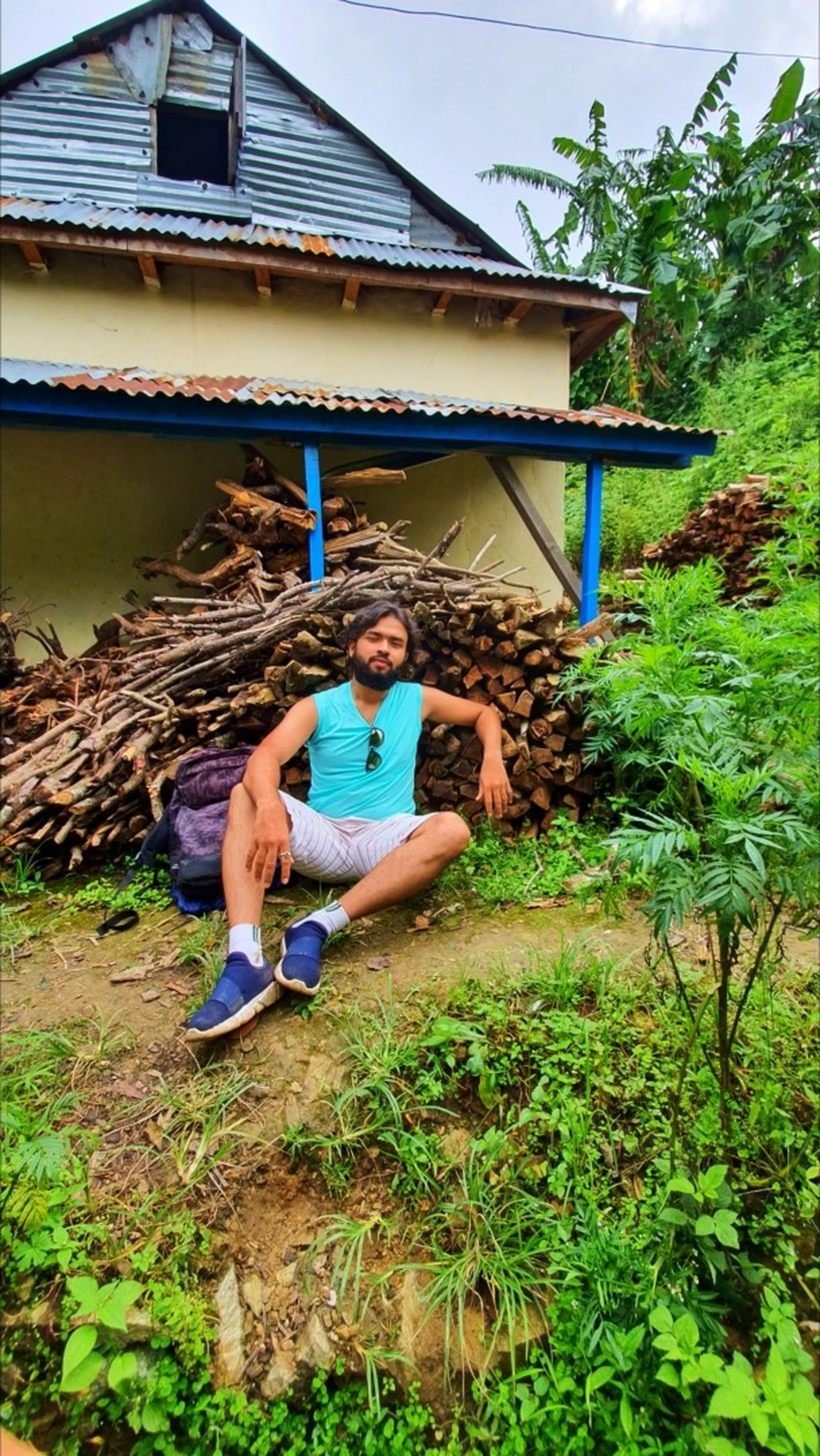 A bearded male trekker in a teal sleeveless tank top, light striped shorts, white socks, and blue sneakers sitting casually cross-legged on the grassy ground in front of a traditional Nepali village house with corrugated tin roof and blue-painted pillars in Manikhel Lalitpur Nepal; a large stack of firewood stored under the porch eave behind him, his trekking backpack resting on the ground, wild green plants growing around - relaxed trekking rest stop Nepal village, Manikhel Lalitpur rural Nepal village trail near Kathmandu