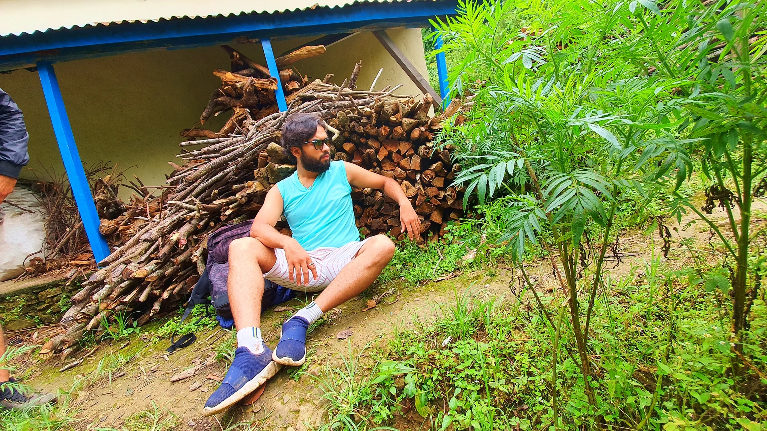 A bearded male trekker in a teal sleeveless tank top, light shorts, and blue sneakers lounging casually on the ground under the blue-pillared porch of a traditional Nepali village house in Manikhel Lalitpur Nepal, resting with his back against a large pile of stacked firewood and legs stretched out; sunglasses hanging from his shirt, backpack on the ground beside him, lush green fern and wild plants in the foreground - authentic Nepal village rest break during trekking, Manikhel Lalitpur rural village near Kathmandu Valley hiking trail