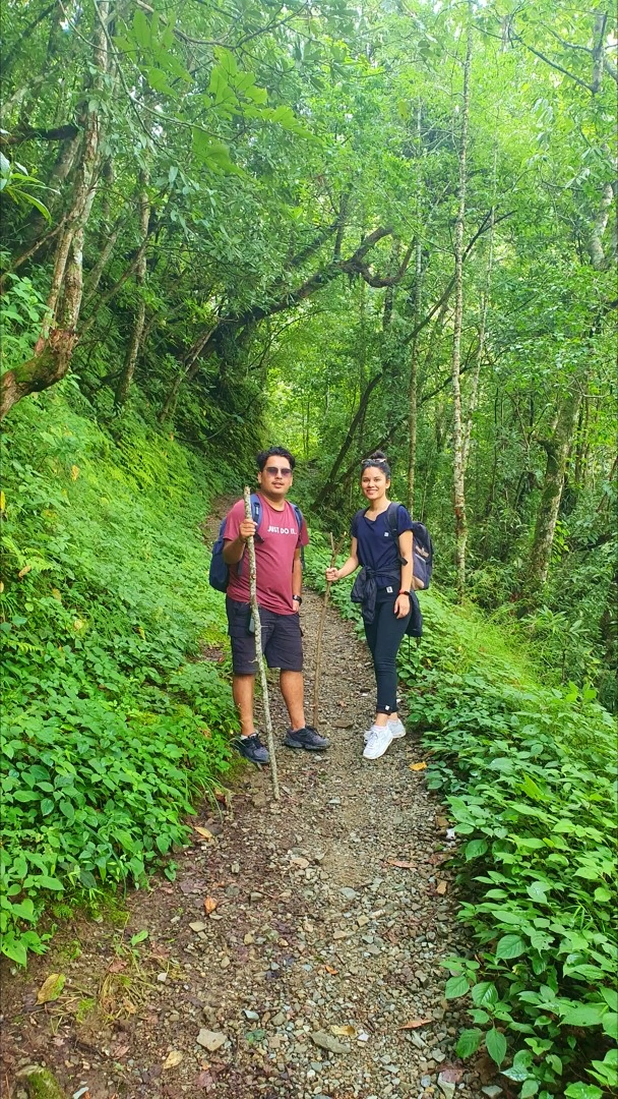 A couple hiking together on a rocky narrow jungle trail in Manikhel Lalitpur Nepal - a man in a maroon t-shirt holding a wooden trekking stick and a woman in a navy blue top with a backpack smiling at the camera; lush green forest canopy arching overhead creating a natural tunnel, dense monsoon vegetation on both sides, ideal Nepal nature trekking trail near Kathmandu Valley