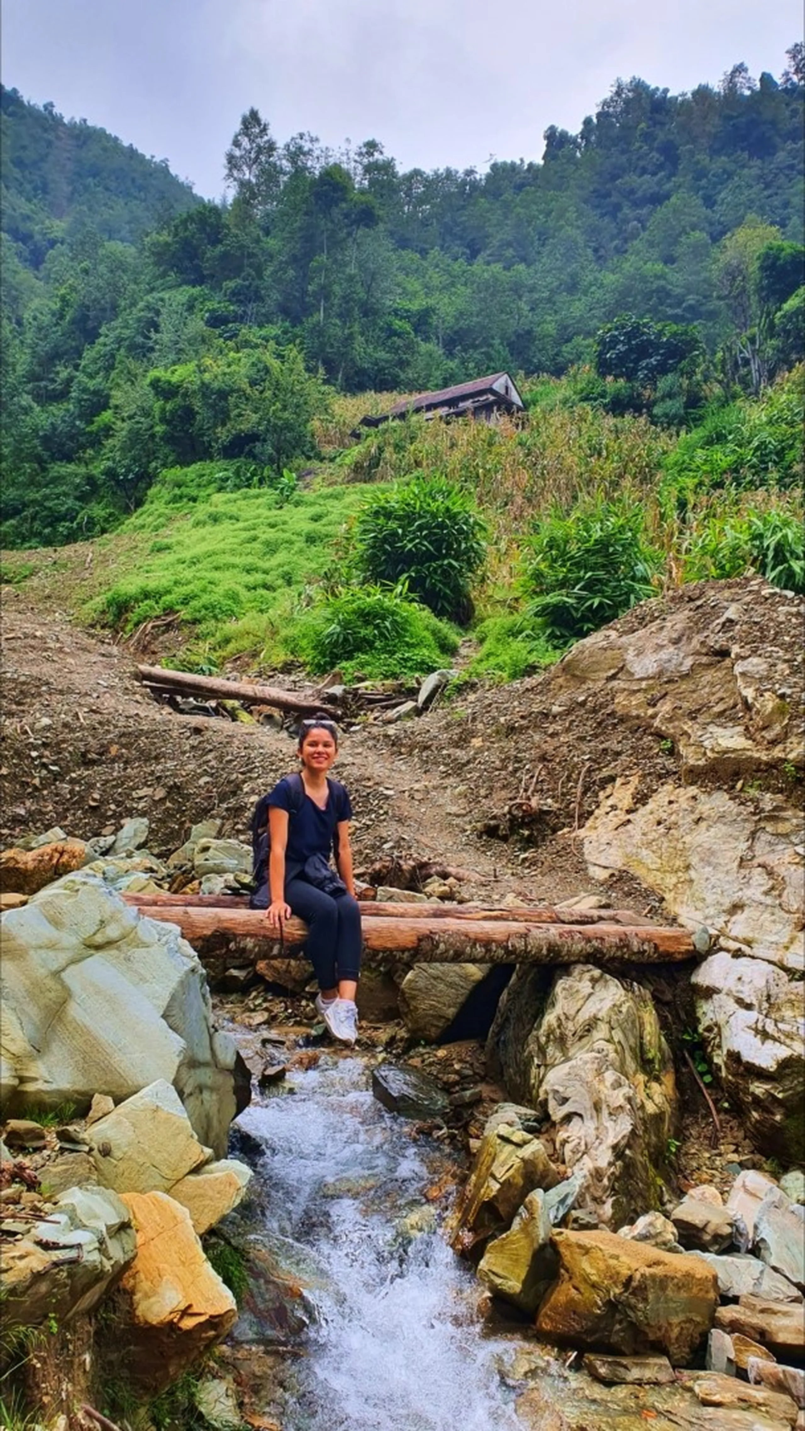 A young woman in all-black trekking outfit sitting relaxed on a rustic wooden log bridge crossing a small rushing mountain stream in Manikhel Lalitpur Nepal; mossy boulders and clear white water flowing beneath her, terraced hillside farmland with bamboo and a traditional tin-roofed Nepali farmhouse visible on the green slope behind her - authentic rural Nepal trekking experience near Kathmandu