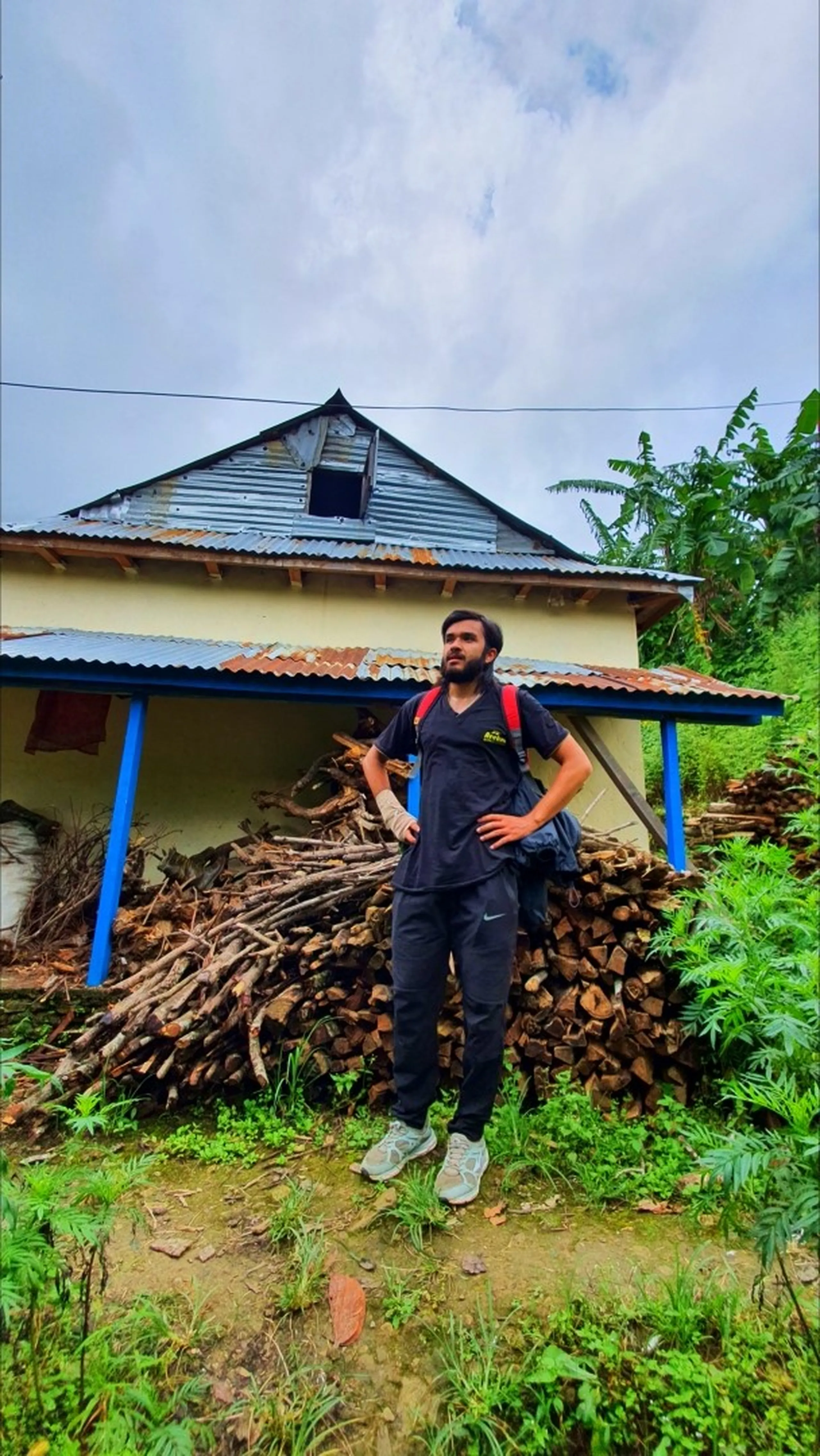 A bearded male hiker in a dark navy scrub-style top with a red backpack standing confidently in front of a traditional Nepali rural house with a corrugated tin roof and blue painted pillars in Manikhel Lalitpur Nepal; a massive pile of stacked firewood logs fills the porch behind him, banana tree and lush greenery on the right - authentic off-the-beaten-path Nepal village trekking scene