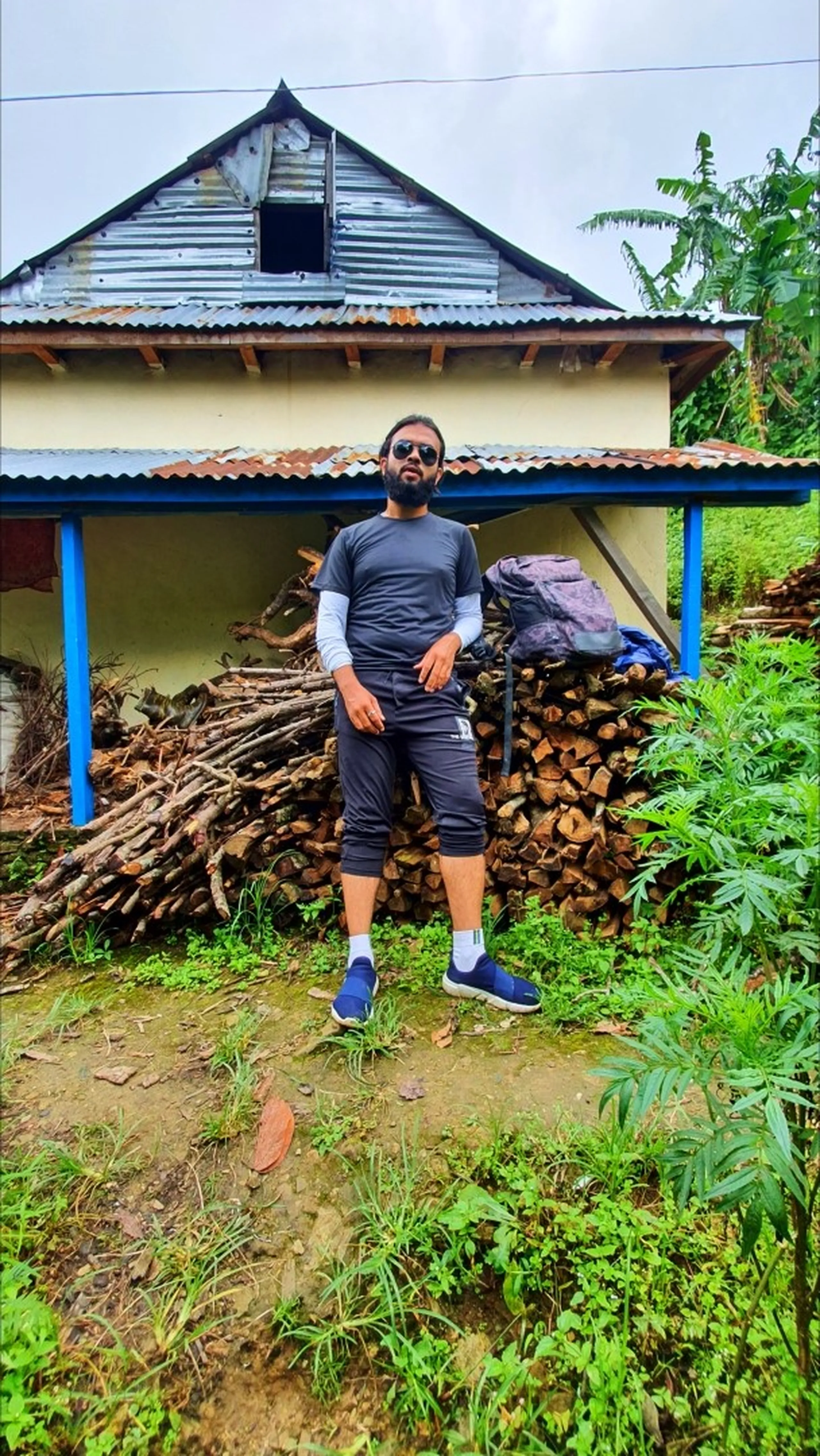 A male trekker wearing a grey athletic shirt layered over long sleeves, dark capri pants, white socks, and blue sneakers with aviator sunglasses standing casually in front of a traditional Nepali village house with a corrugated metal roof in Manikhel Lalitpur Nepal; large stacked firewood pile behind him, wild green plants around - real Nepal village trekking atmosphere on the way to Simba Falls Lalitpur