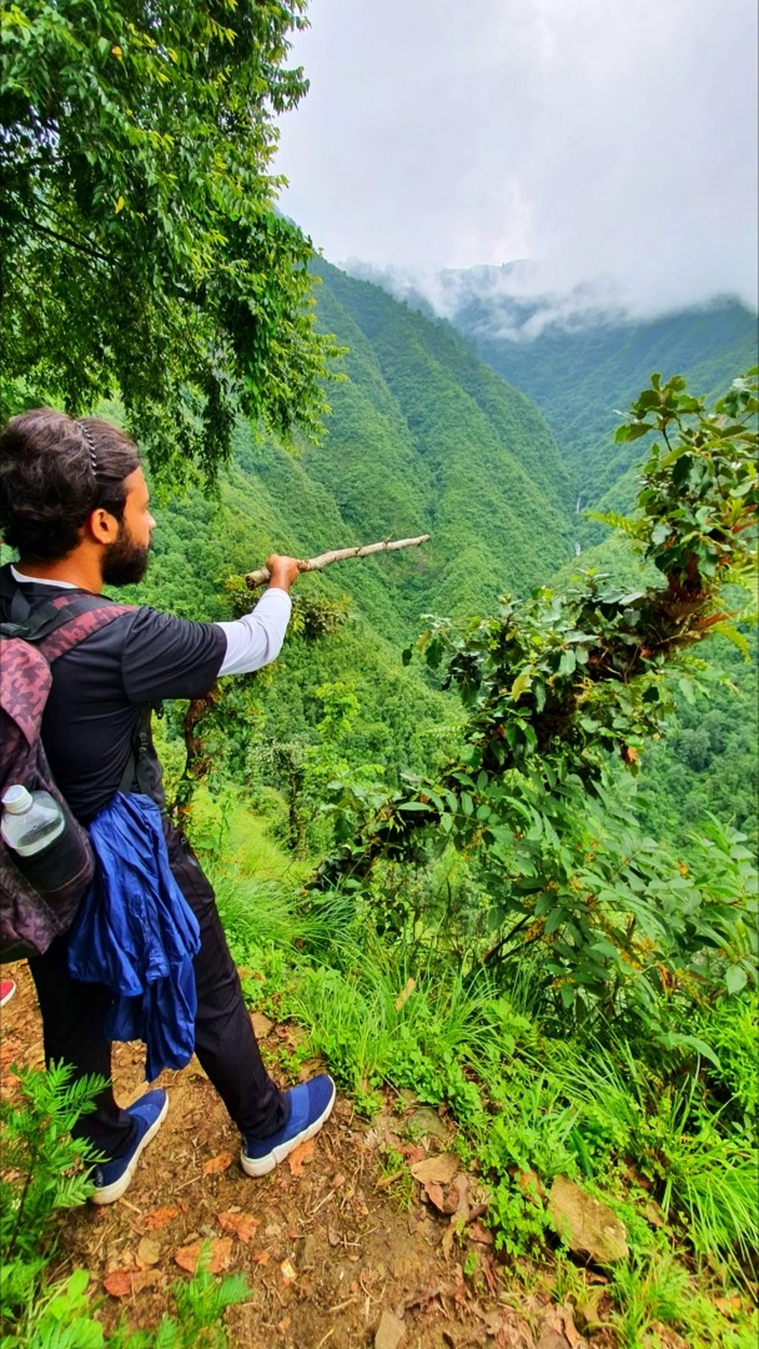 A male trekker with a backpack and trekking stick seen from behind pointing dramatically with his stick toward a breathtaking deep green gorge in Manikhel Lalitpur Nepal; steep jungle-covered Himalayan foothills plunging into a dramatic V-shaped valley with a thin white waterfall cascading down the far cliffs through monsoon mist and low clouds - epic hidden waterfall viewpoint on the Simba Falls trail near Kathmandu Nepal