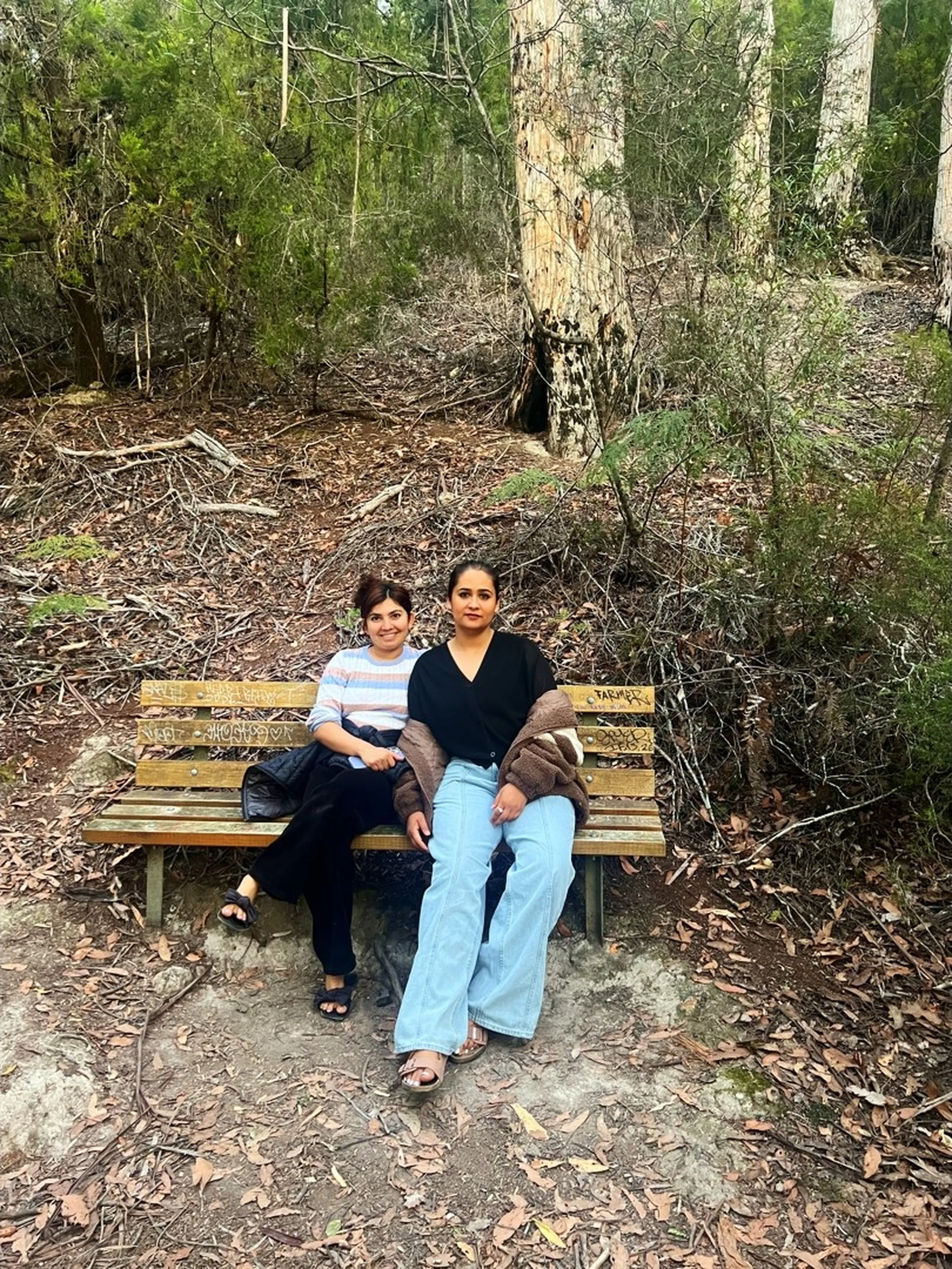 Two young women sit side by side on a weathered wooden park bench in the middle of a dense Australian bushland setting. The bench has graffiti scratched into its backrest, including the word "FARMER." The ground around them is covered in dry brown leaf litter, fallen twigs, and scattered bark. Behind them, the bush is thick and layered - tall eucalyptus trees with pale, peeling bark rise in the background, one with a large hollow at its base. The woman on the left is shorter, wearing a light blue and pink horizontal-striped top with black pants and black sandals, a dark bag beside her. The woman on the right is taller, wearing a black V-neck top, light blue wide-leg jeans, and beige sandals, with a brown fluffy jacket draped over her lap. Both are looking directly at the camera with relaxed, natural smiles. The atmosphere is cool, shaded, and quietly wild.