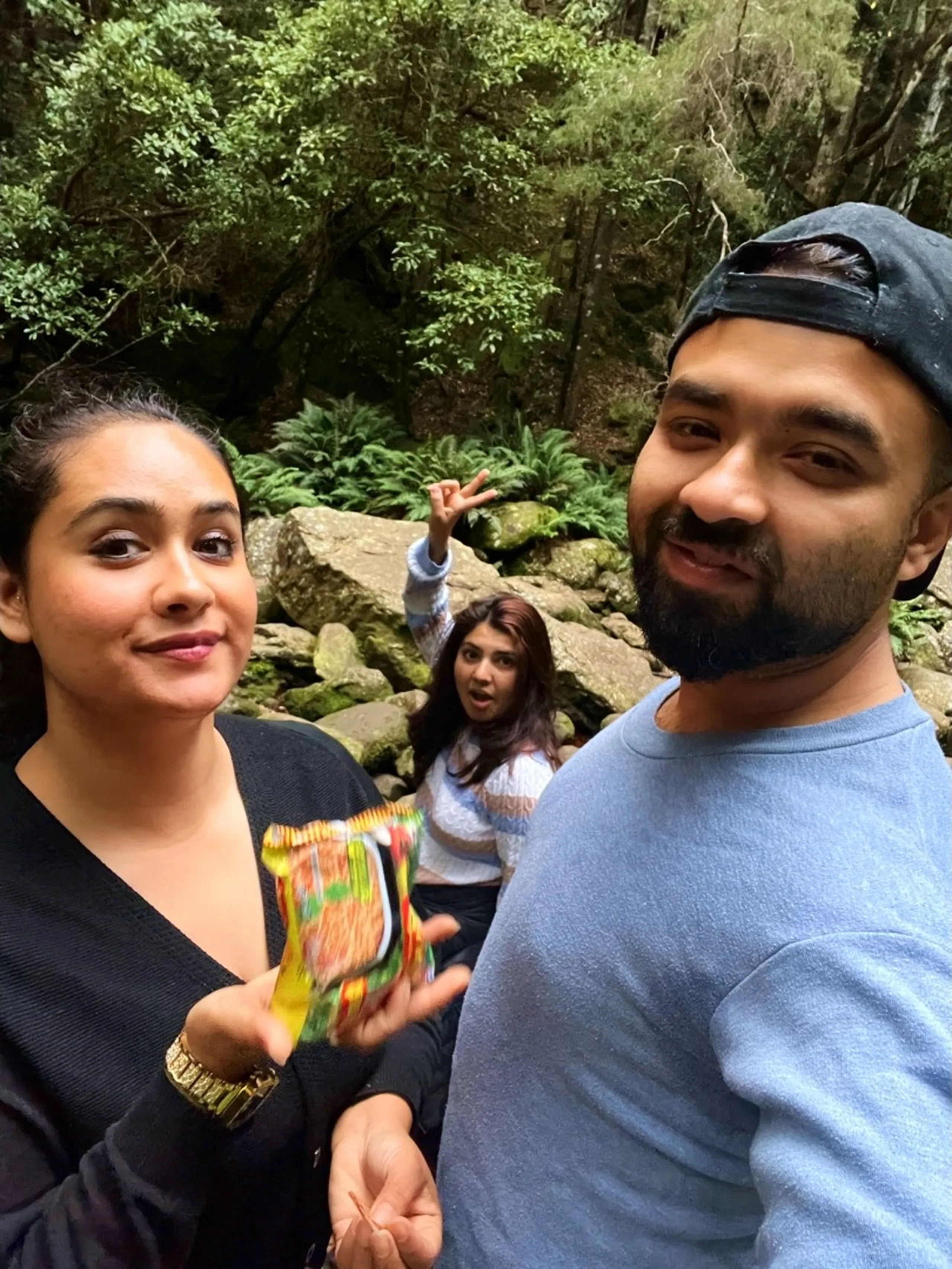 A playful, candid selfie taken in the forest near the waterfall area. In the foreground, a young woman in a black top holds a colorful object - what appears to be a brightly colored snack wrapper or toy - toward the camera, with a mischievous half-smile and wide eyes. Beside her on the right, the bearded man in the blue sweater and backwards black cap grins at the camera. In the background, slightly out of focus but clearly photo-bombing deliberately, a third woman in the striped sweater sits on a boulder with her mouth open wide in a dramatic expression and one hand raised making a peace sign. Dark green ferns and mossy rocks fill the background. The mood is silly, spontaneous, and full of laughter.