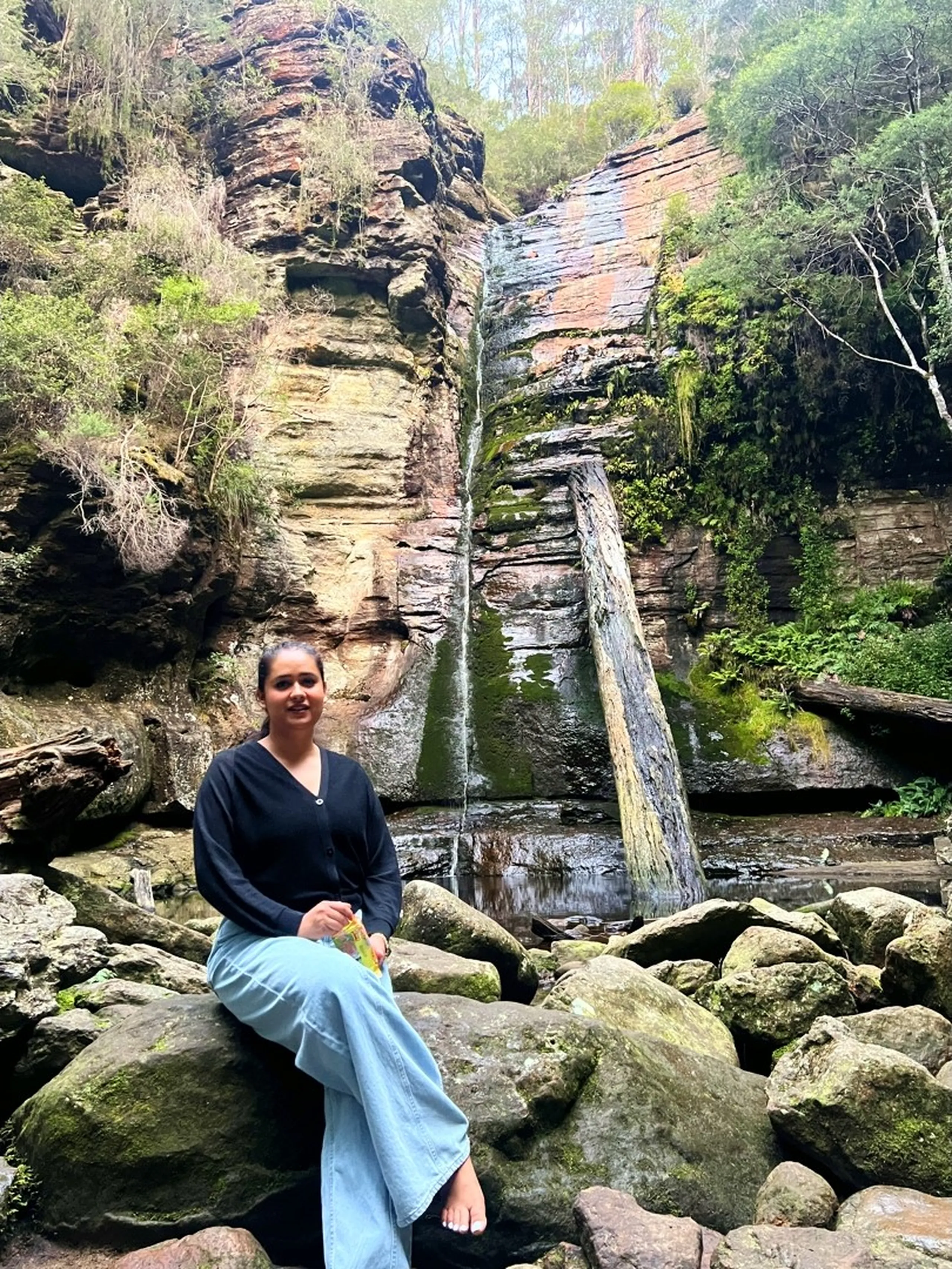 A full-body portrait of a woman sitting barefoot on a large rounded moss-speckled boulder at the waterfall base. She wears a black long-sleeve V-neck top and light blue wide-leg jeans, one knee bent upward. Her bare feet rest on the rock. She holds what appears to be a small snack or wrapper in her hands on her lap and smiles gently at the camera. The full waterfall is visible behind her - the narrow silver stream falling the entire height of the sandstone cliff, the diagonal fallen log, and the still pool at the base. The cliff walls are dramatically lit with warm afternoon light, revealing deep orange and rusty red mineral bands in the rock. The ferns and green vegetation add lushness to the sides. The woman appears entirely at ease, as if she belongs to the landscape.