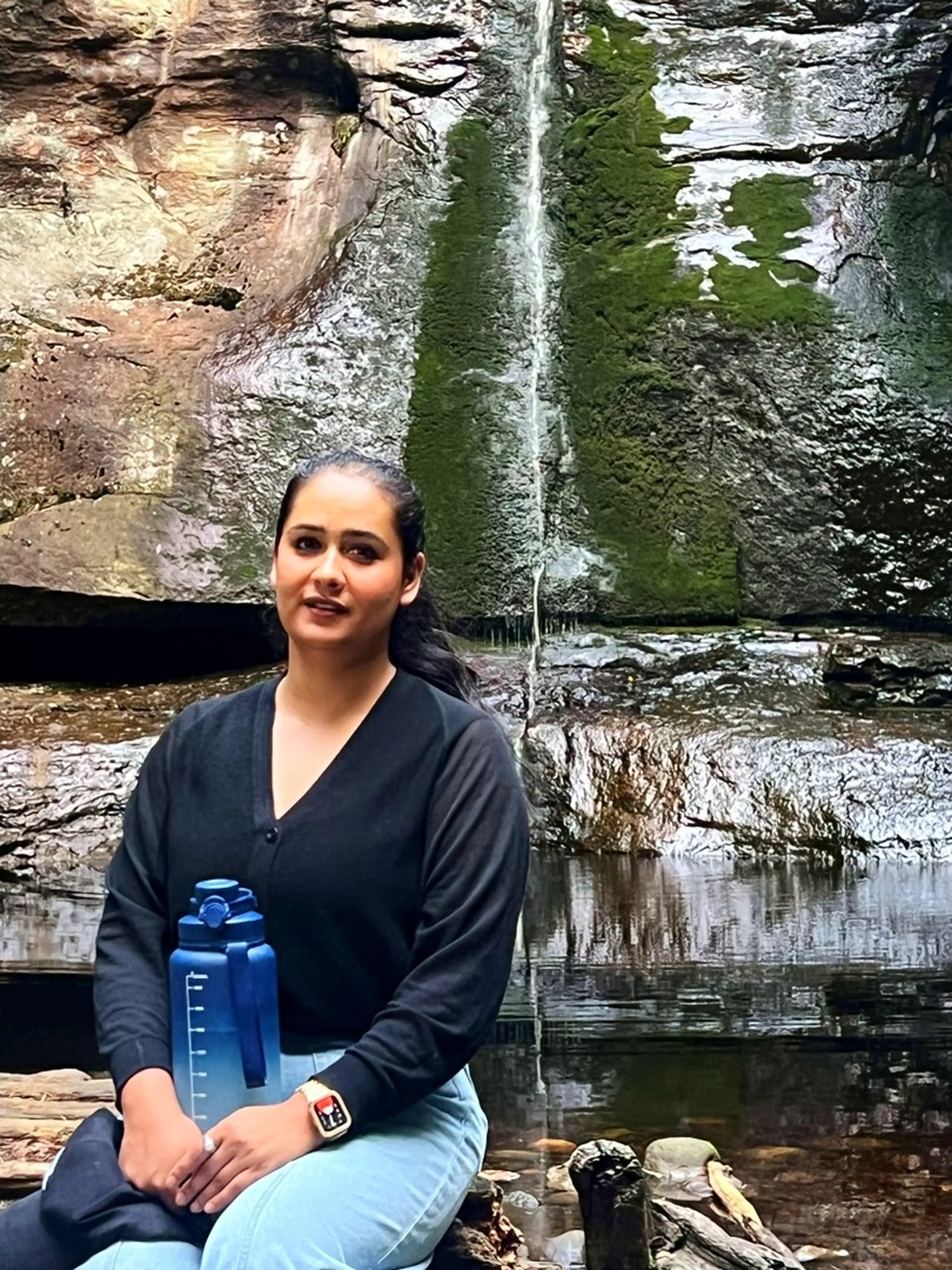 A close-up portrait of a young woman sitting at the base of the waterfall, directly in front of the lower section of the cliff face. The sandstone wall behind her is dark, wet, and covered in patches of vivid green moss. A thin stream of water falls vertically just behind her head and cascades onto a flat wet ledge, creating a shallow reflective pool. She wears a black V-neck cardigan top and light blue jeans, holds a blue gradient water bottle with measurement markings in her right hand, and wears a rose-gold Apple Watch on her left wrist. Her dark hair is pulled back and she looks at the camera with a calm, soft smile. The proximity to the waterfall and the mossy ancient rock give the portrait a moody, intimate, almost mystical quality.