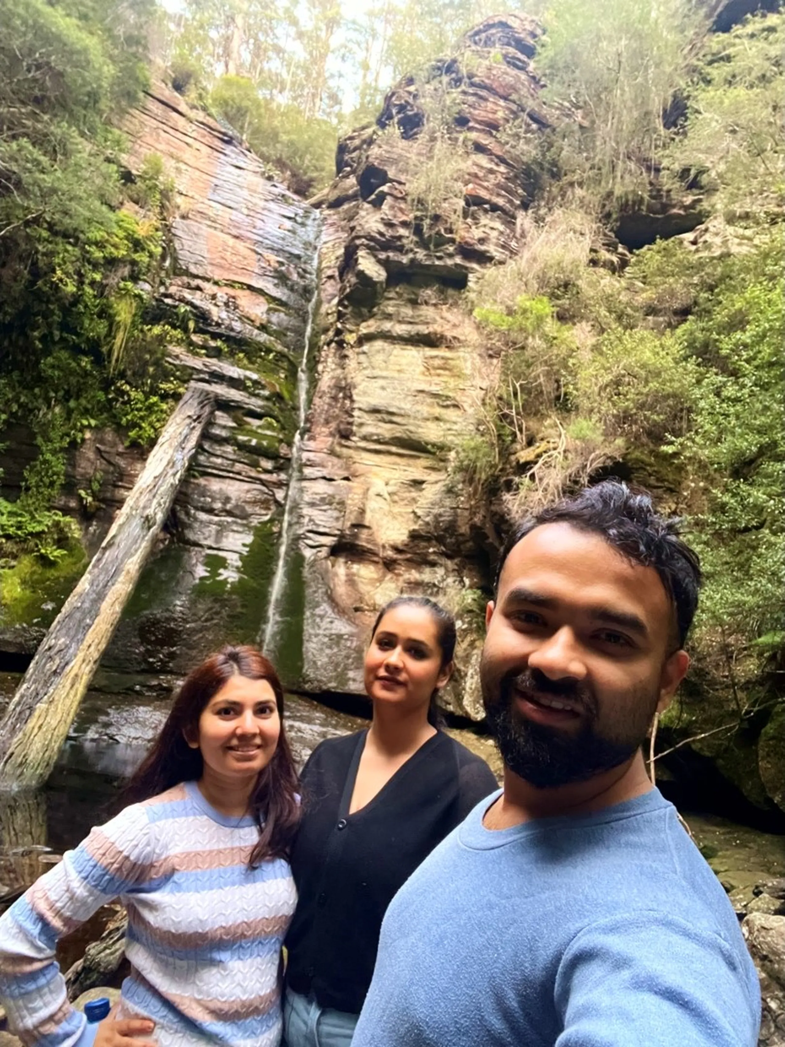 A selfie of three people standing together at the base of the same waterfall gorge, with the towering sandstone cliff and thin waterfall filling the background. The man on the right - bearded, wearing a blue crew-neck sweater - takes the photo. To his left stands a woman in a black top with a warm smile, and behind her in the center is another woman in the striped patterned sweater, slightly further back. The cliff face rises dramatically behind them with its layered orange-rust sandstone, green moss, and vegetation. The diagonal fallen log is visible on the left side of the frame. The three are close together, expressions warm and happy. The image has the feeling of a triumphant end-of-hike moment.