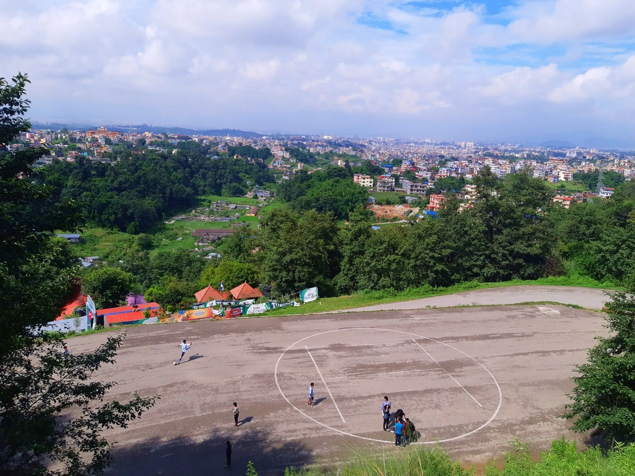 Aerial panoramic view of Kathmandu Valley from Kapan hilltop with children playing football on a dirt ground, lush green forested hills, dense urban cityscape stretching to the horizon under dramatic blue sky with white clouds - Kathmandu day hike viewpoint Nepal