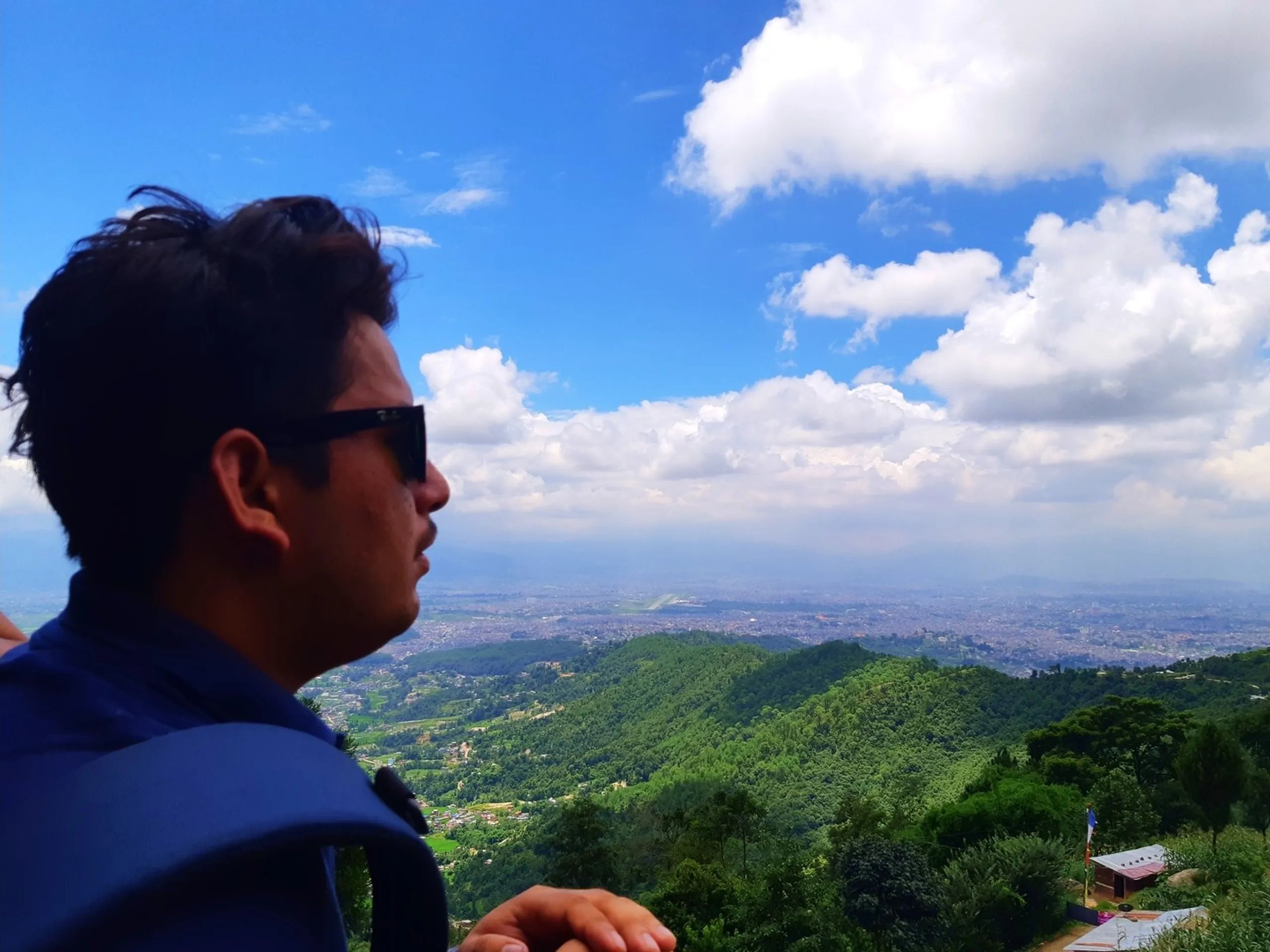 Side profile of a young male traveler wearing dark sunglasses gazing contemplatively at a vast aerial panoramic view of Kathmandu Valley, rolling green forested hills and dense city spread below, bright blue sky and fluffy white clouds - Kathmandu Valley viewpoint trekking Nepal best views