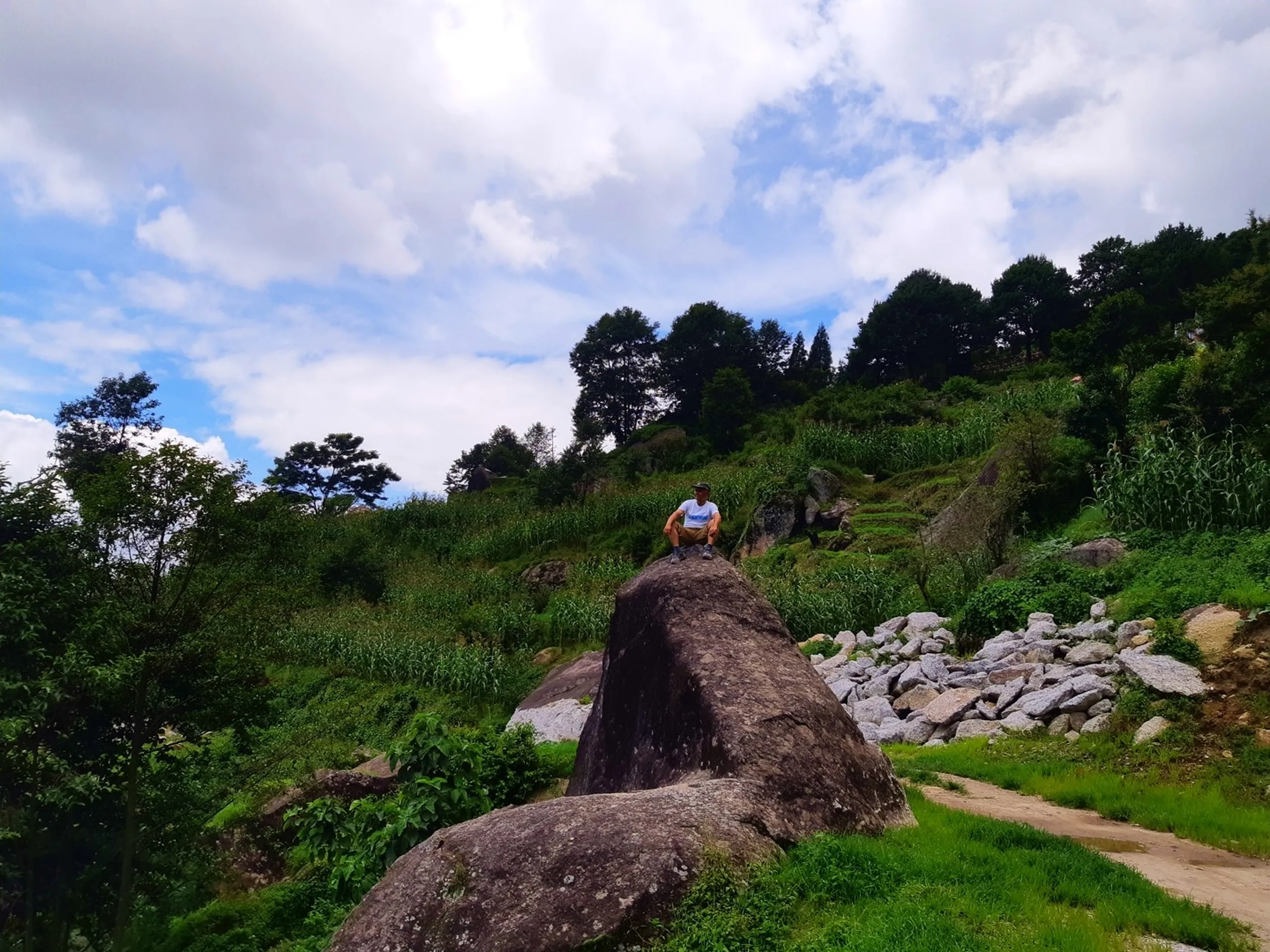 Male hiker in white t-shirt and cap sitting perched confidently on top of a large granite boulder on a hillside trail, lush green terraced farmland with maize crops and scattered pine trees climbing the slope behind, cloudy sky - Nepal hillside trekking boulder viewpoint Kapan Sundarijal