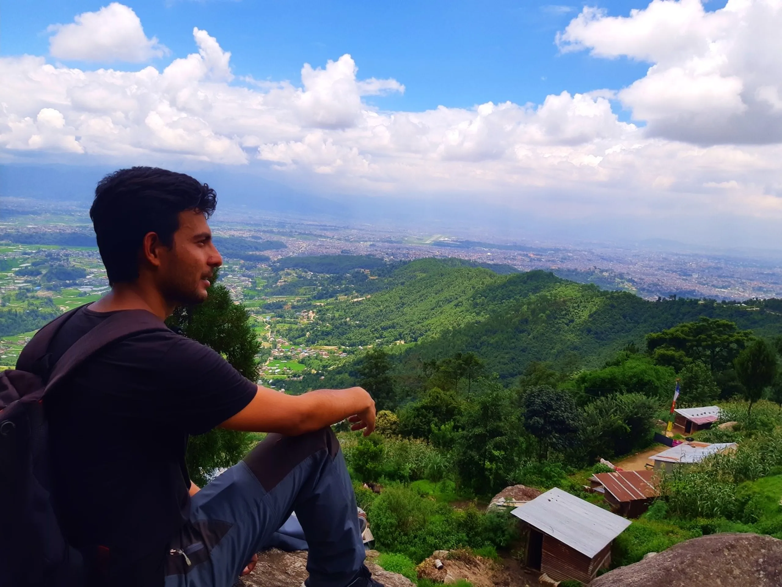 Young male trekker sitting relaxed on rocky outcrop with a spectacular bird's-eye view of Kathmandu Valley spread wide below, green forested ridges in midground, small Buddhist shrine with prayer flags visible, blue sky with white clouds - Kathmandu Valley panoramic view Nepal best trekking viewpoint