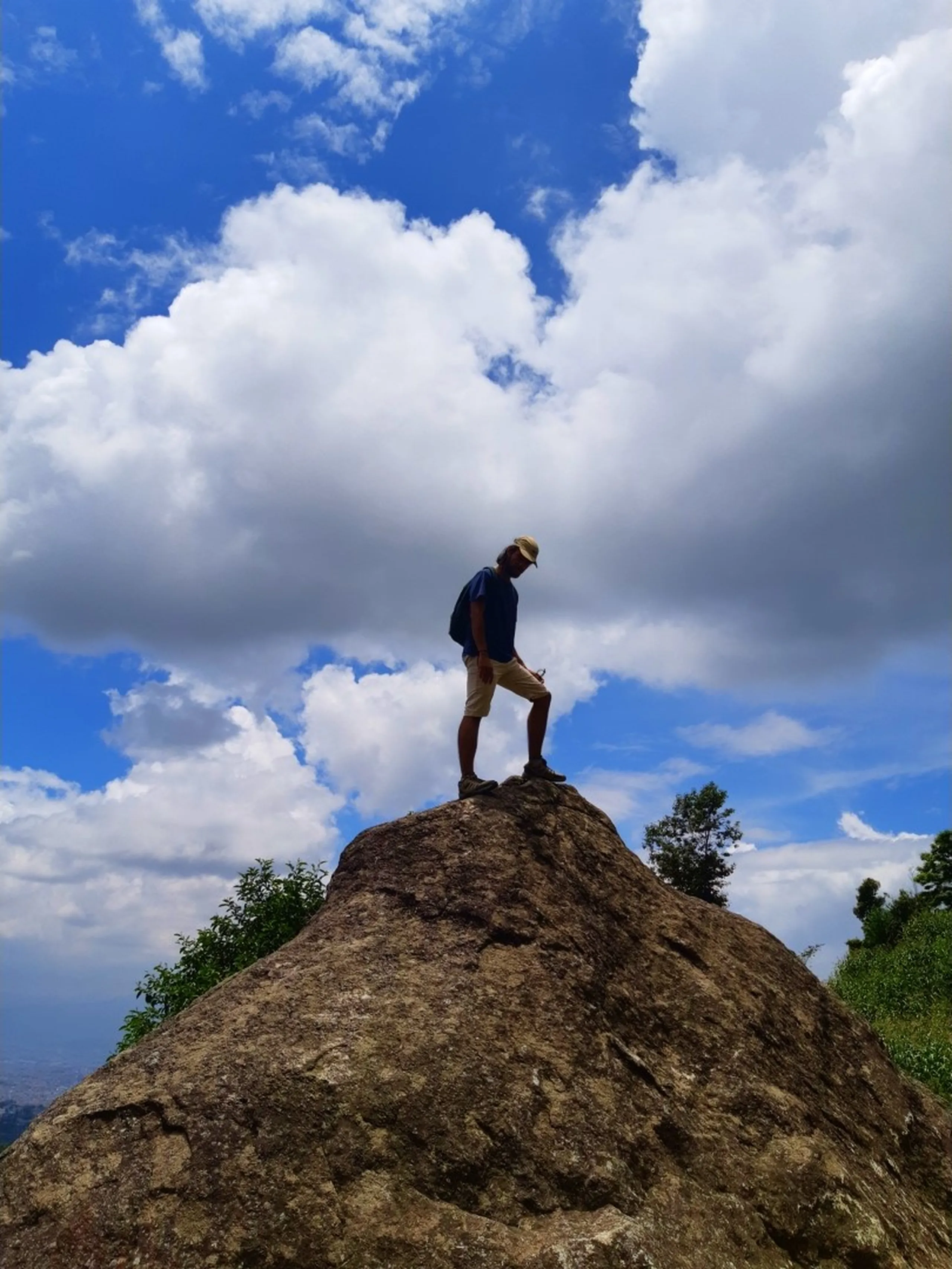 Dramatic low-angle silhouette of a lone hiker standing triumphantly on top of a large rounded granite boulder against a brilliant blue sky filled with dramatic white clouds, green shrubs framing the sides - Nepal hilltop trekking adventure solo hiker dramatic sky