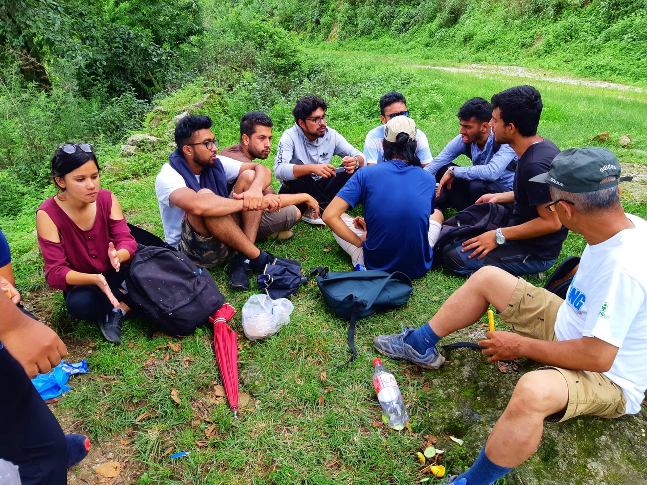 Large group of young Nepali hikers and friends sitting together in a casual circle on green grass beside a forest trail during a trekking break, backpacks scattered around, lively conversation and group bonding - Nepal group trekking friends day hike Sundarijal Kapan trail