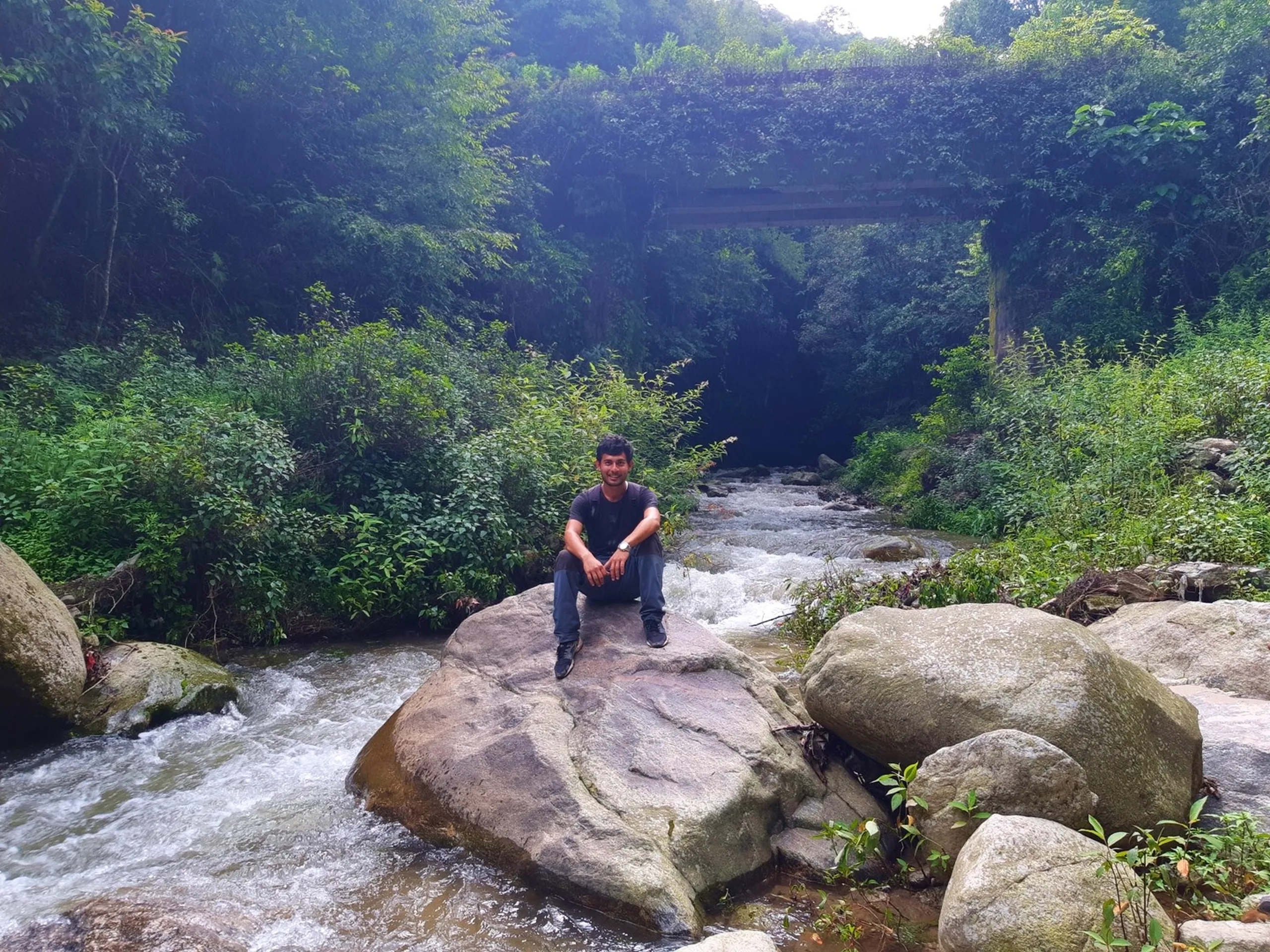 Happy male hiker sitting cross-legged on a large midstream boulder in a rushing mountain river with white water rapids flowing around him, lush green jungle vegetation and a concrete bridge visible in the background - Sundarijal river trekking Nepal mountain stream hike