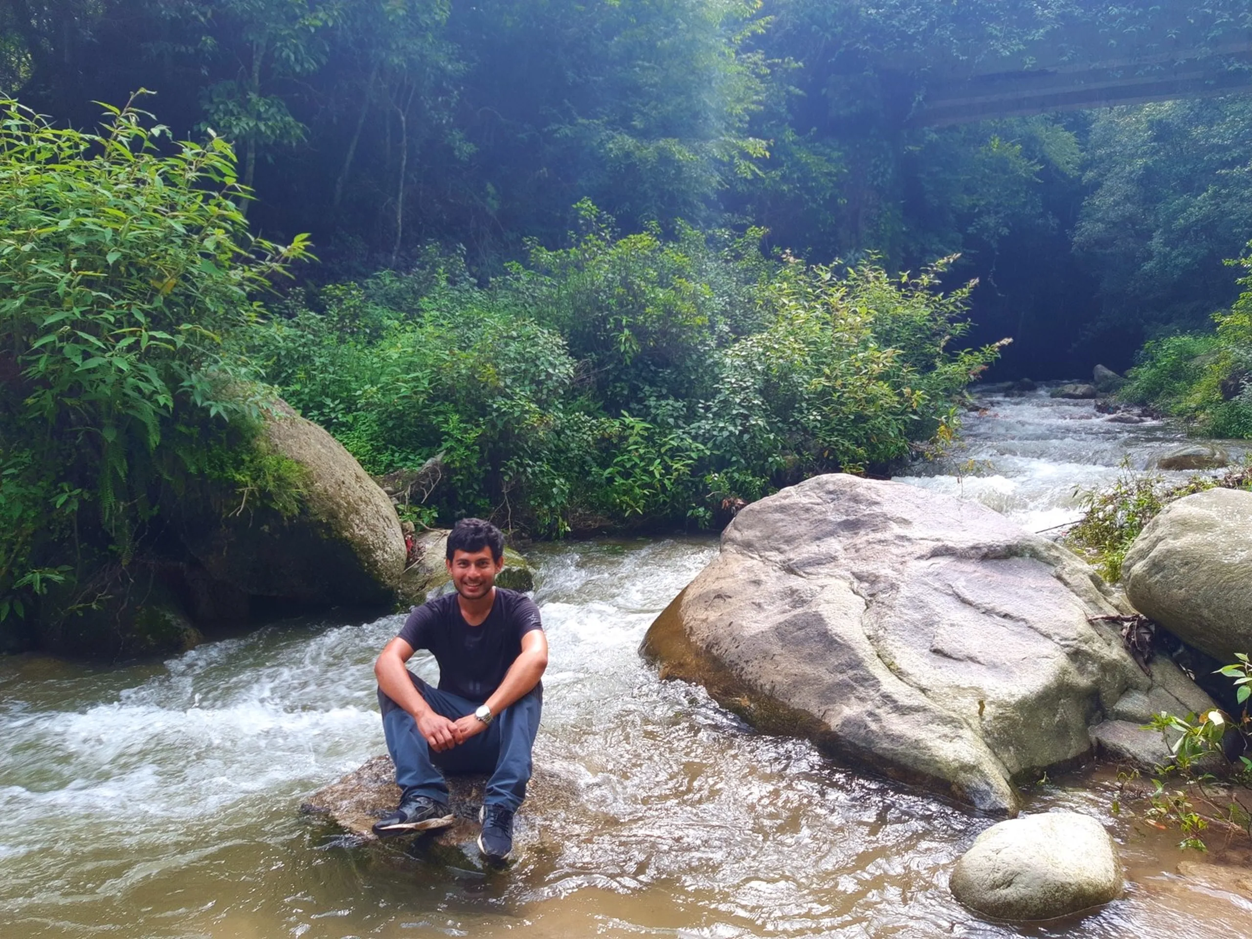 Smiling young male traveler crouching on a flat rock in the middle of a fast-flowing rocky mountain stream, feet close to the rushing water, surrounded by dense dark green monsoon forest and large boulders - Nepal mountain river stream trekking Sundarijal Kapan hike
