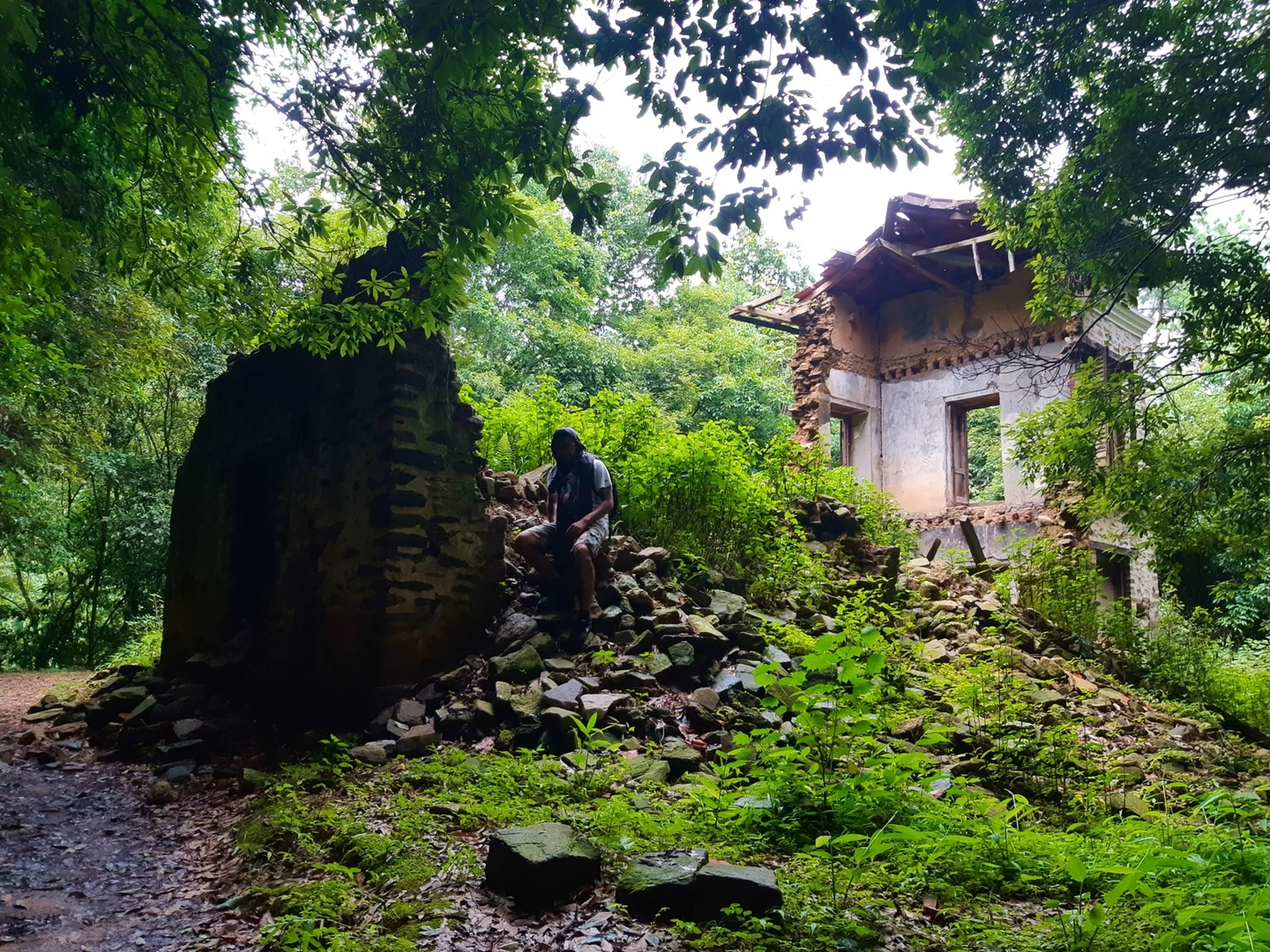Male hiker sitting on a pile of crumbling stone and brick rubble of an earthquake-destroyed abandoned Nepali house slowly being reclaimed by lush green jungle, partially standing stone wall and deteriorating white plastered structure visible - Nepal 2015 earthquake ruins abandoned village Sundarijal trekking heritage