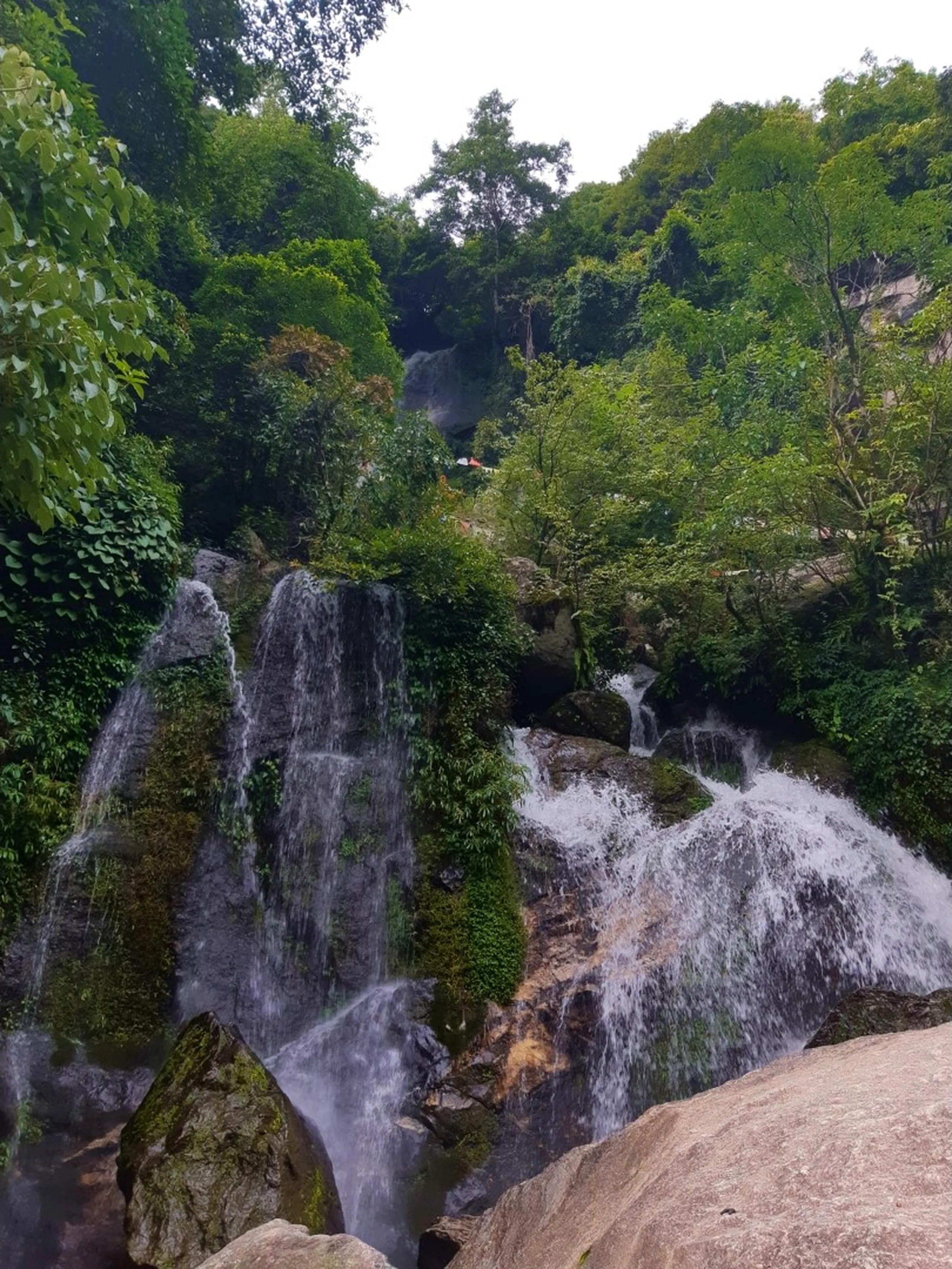 Multi-tiered Sundarijal waterfall cascading powerfully over moss-covered rocks through dense tropical green jungle, white silky water splitting into multiple streams over boulders, lush monsoon vegetation framing both sides, another waterfall visible higher up in the background - Sundarijal waterfall Nepal best waterfall near Kathmandu trekking destination