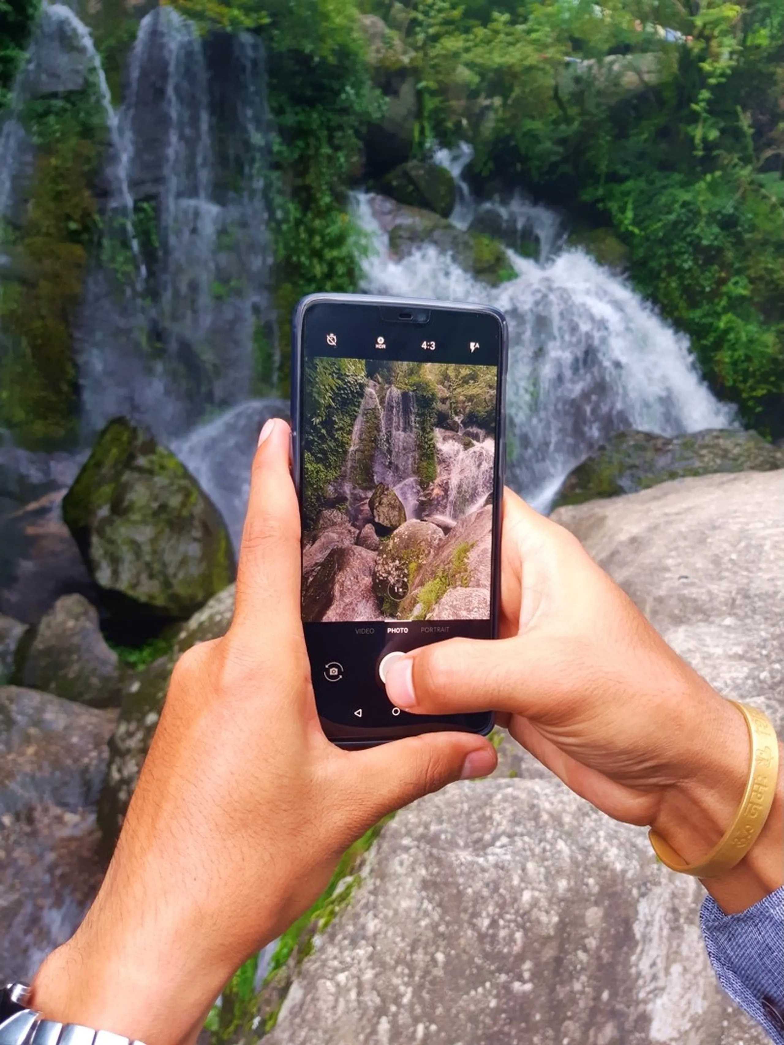Close-up of male hiker's hands holding a smartphone capturing a photo of Sundarijal waterfall rushing over moss-covered rocks, the waterfall reflected live on the phone screen creating a picture-within-a-picture effect, gold bracelet and watch visible on wrists - Nepal travel photography waterfall smartphone hiking Sundarijal